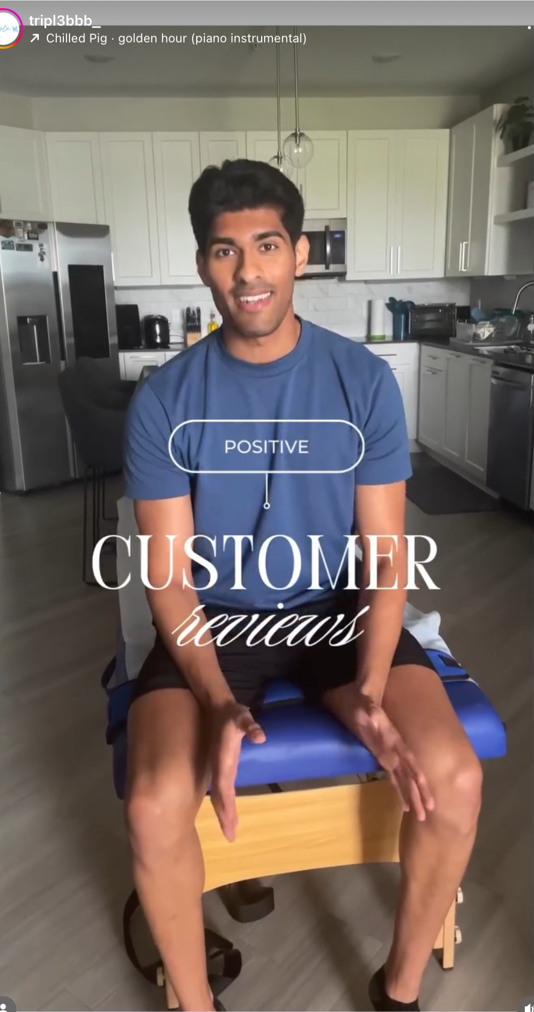 Young man sitting on a chair in a kitchen, smiling at the camera, wearing a blue t-shirt and shorts.