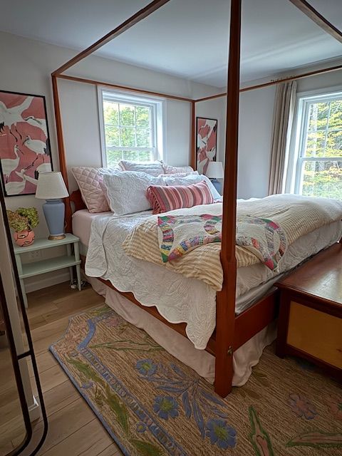 Bedroom with a four-poster bed, white bedding, colorful pillows, and two windows with natural light.