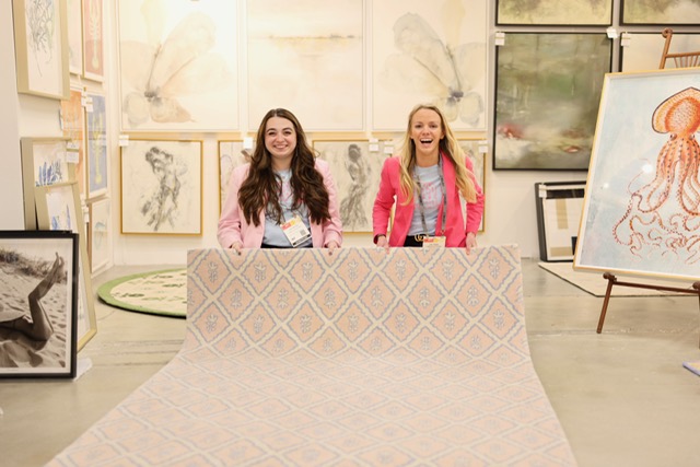 Two women stand behind a large table in an art gallery, smiling, with artwork displayed on walls and easels around them.
