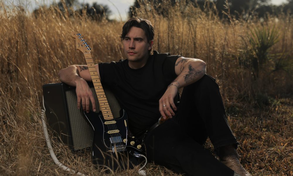 Person sitting outdoors with a guitar and amplifier, surrounded by tall grass and trees in the background.