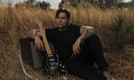 Person sitting outdoors with a guitar and amplifier, surrounded by tall grass and trees in the background.