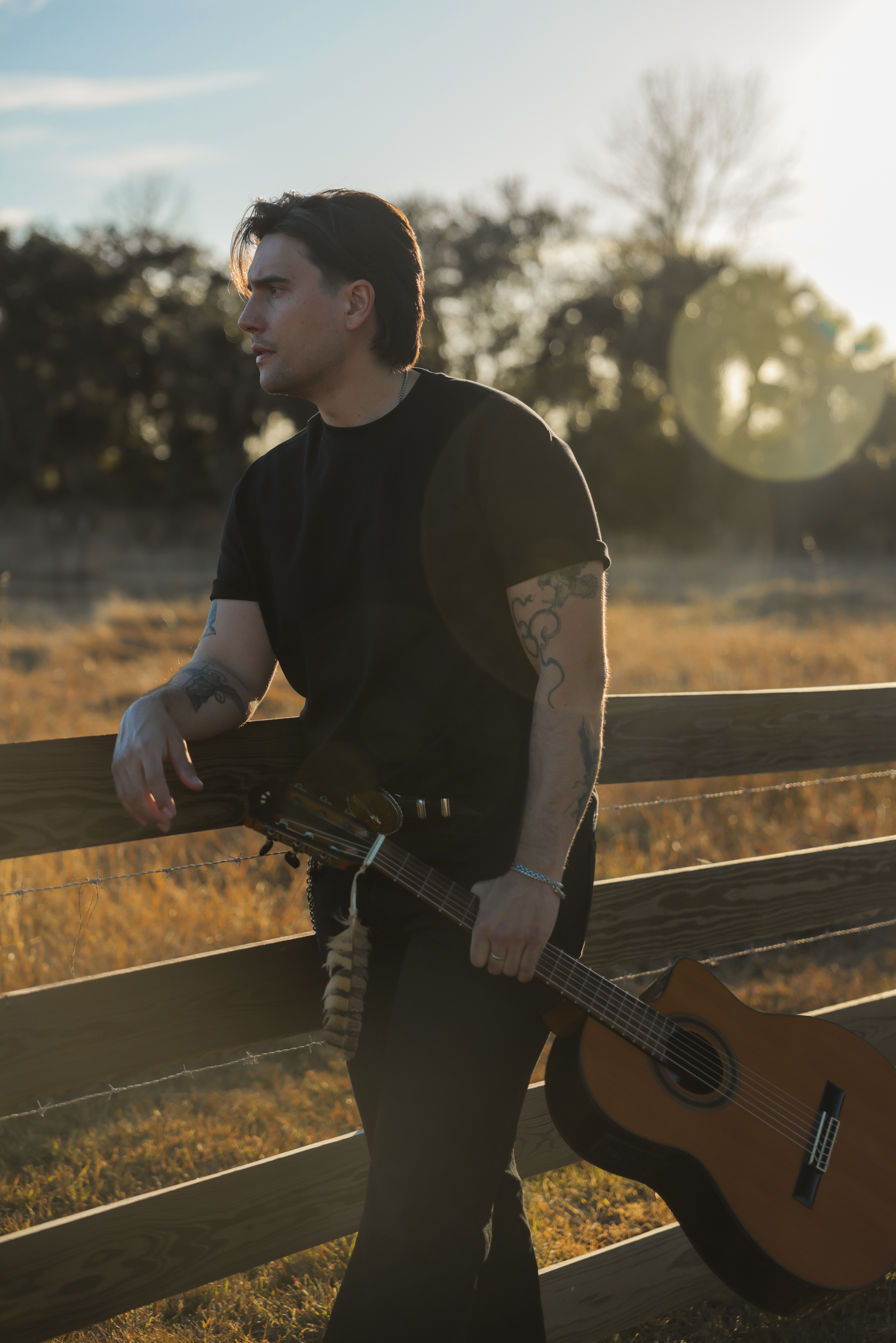 Young man with dark hair and tattoos standing outdoors holding a guitar, with trees and sunlight in background.