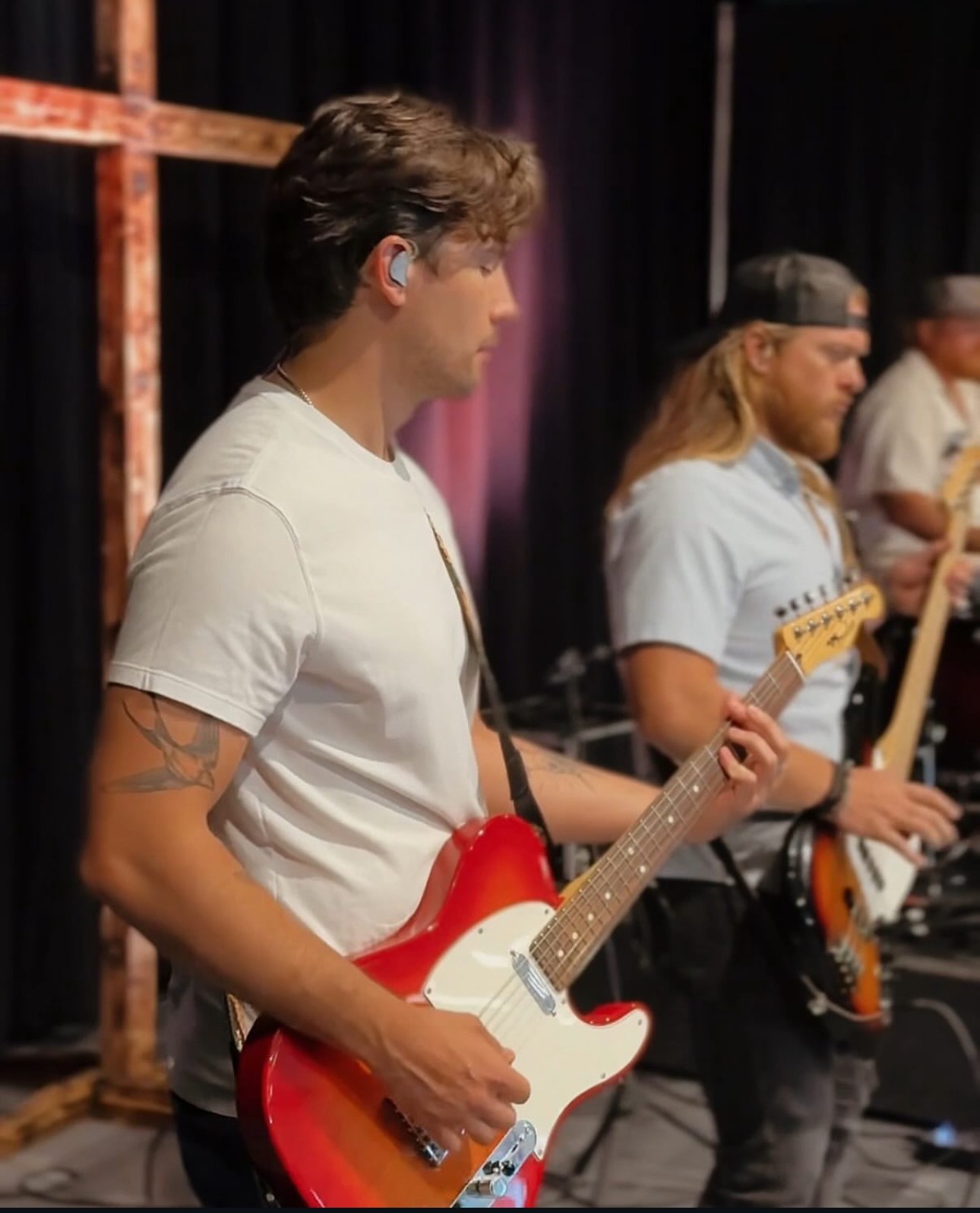 Young man playing a red electric guitar on stage with other musicians in the background.
