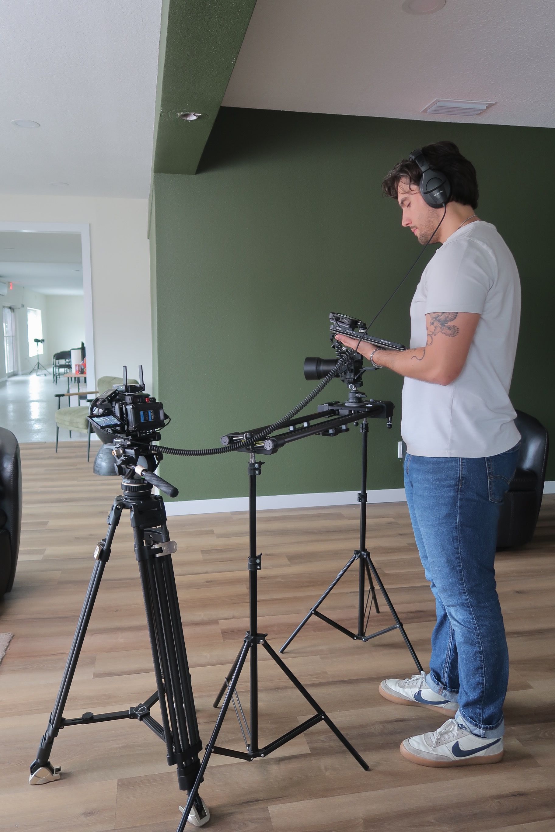 Man with headphones operating camera and tripod in a room with green wall and wooden floor.