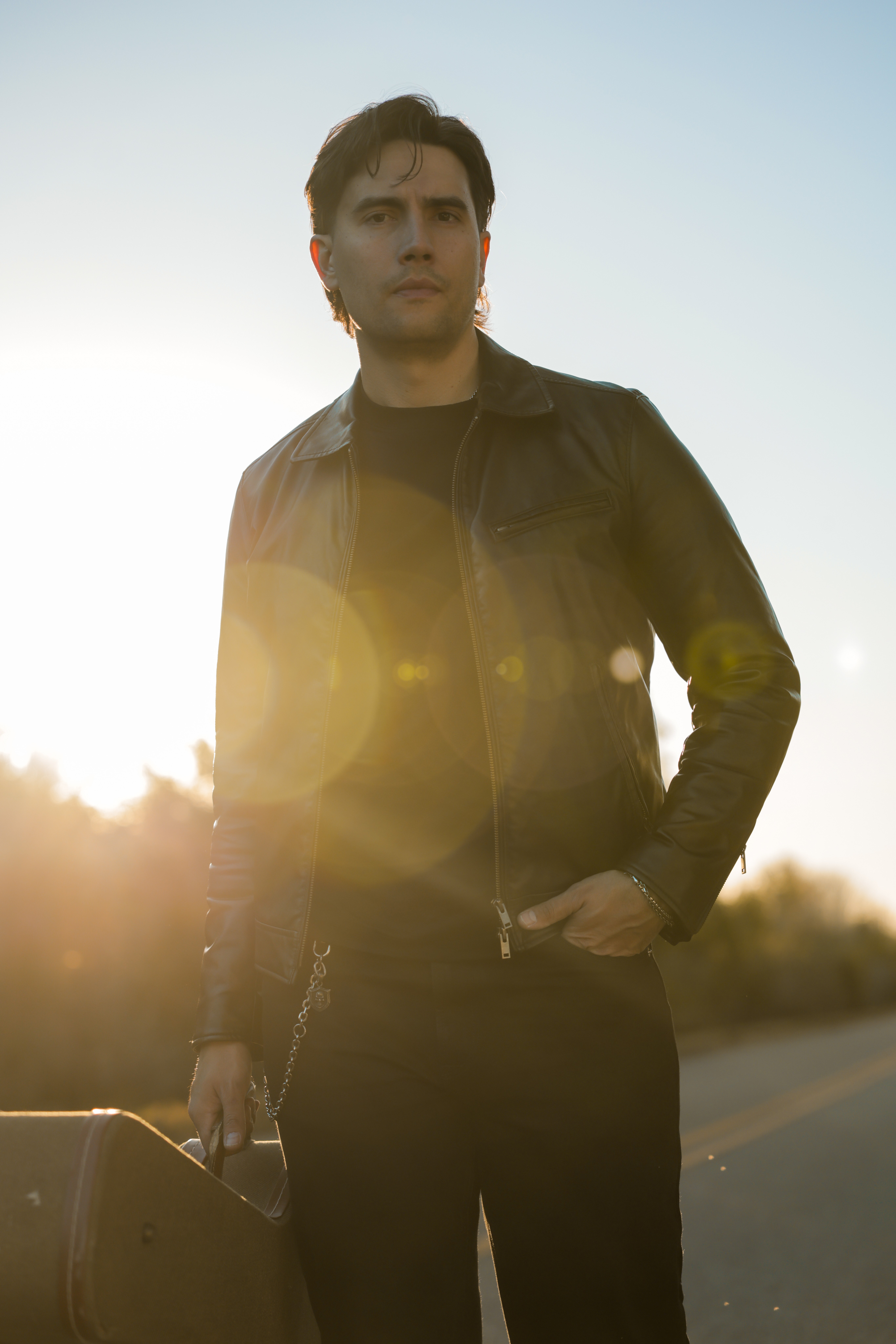 Young man standing outdoors with sunlight behind him, wearing a black leather jacket and holding a bag.