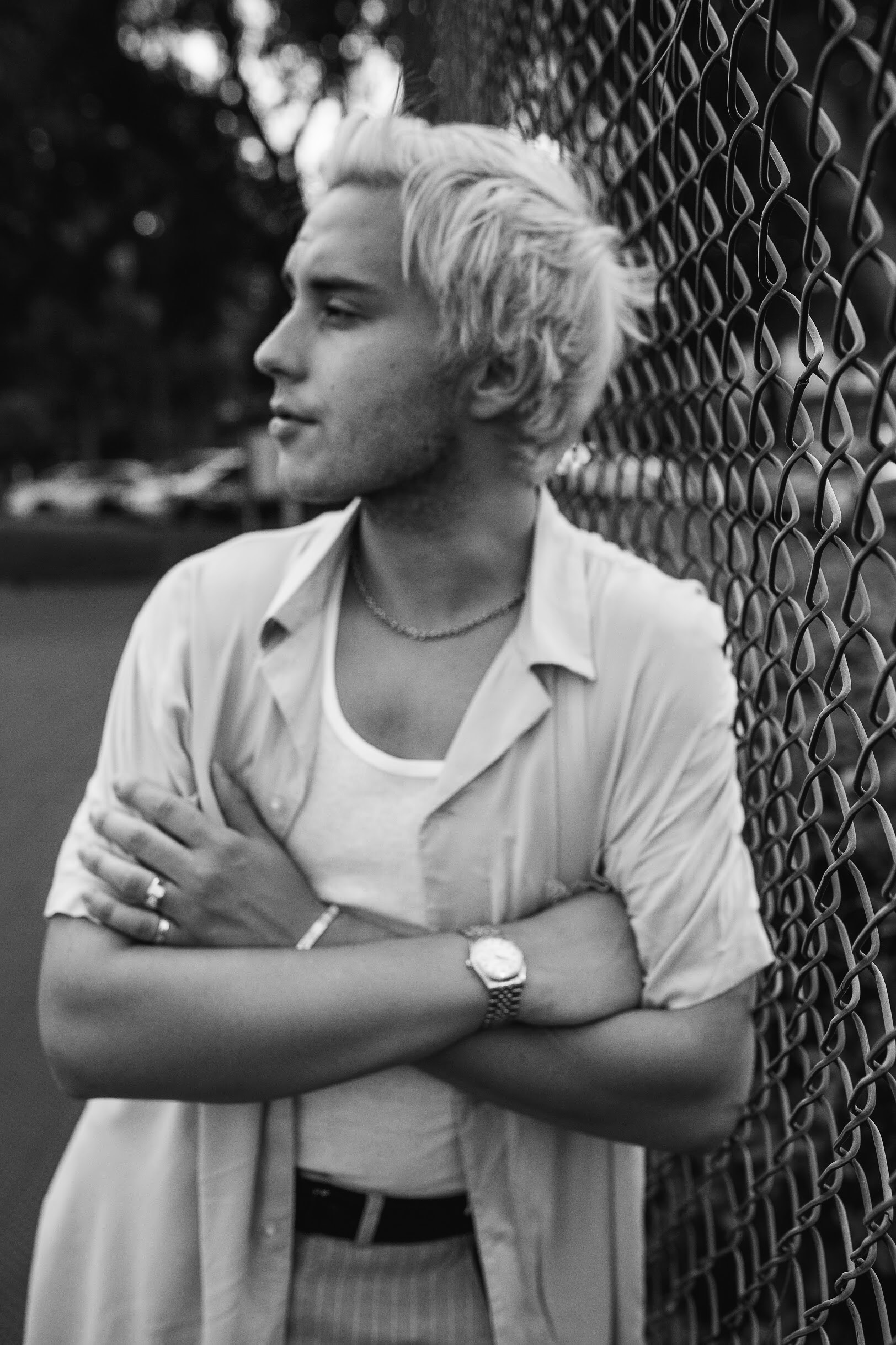 Young person with short, light-colored hair standing next to a chain-link fence, arms crossed, looking to the side.