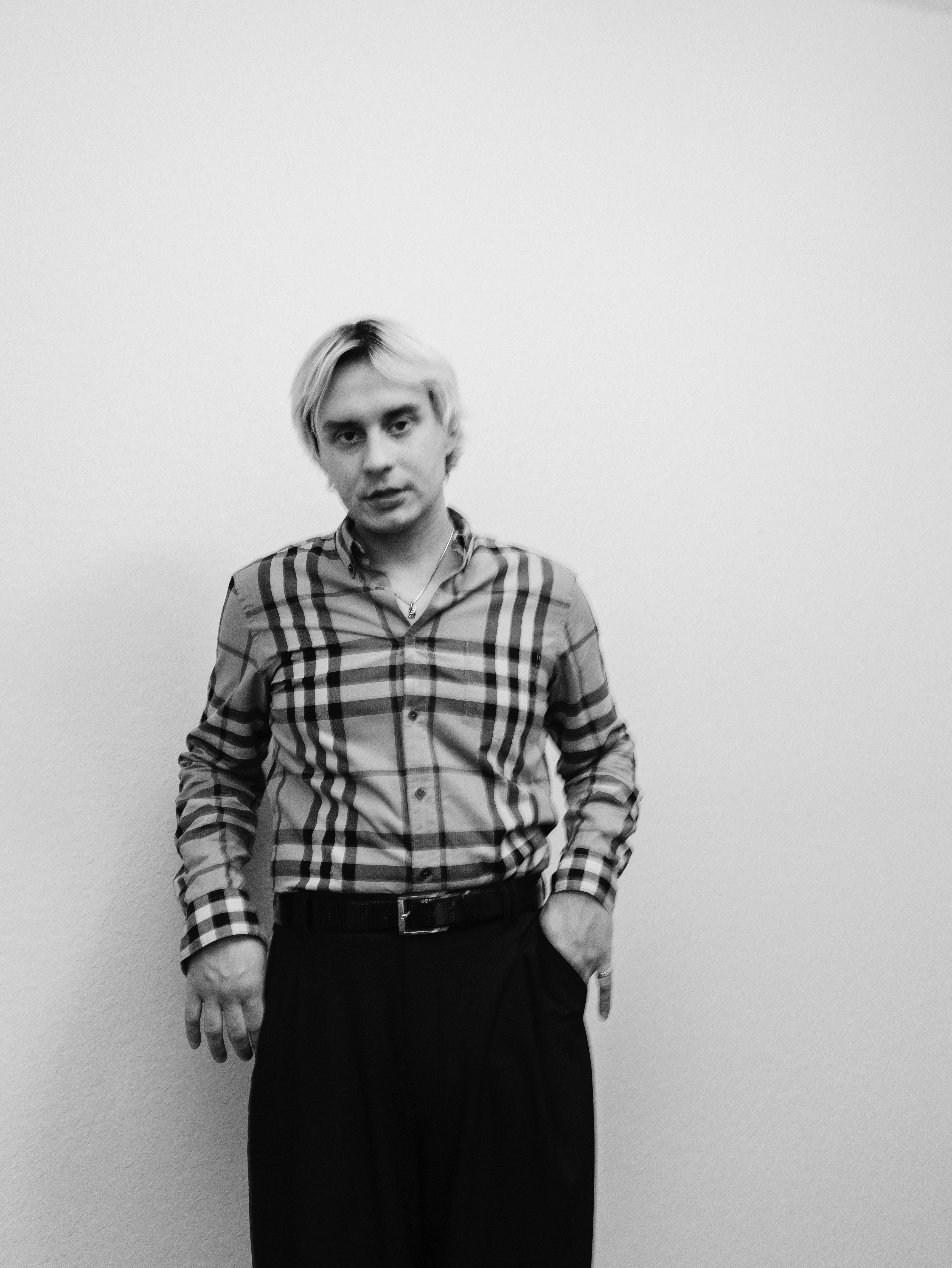 Person with short hair wearing a plaid shirt and dark pants, standing against a plain wall.