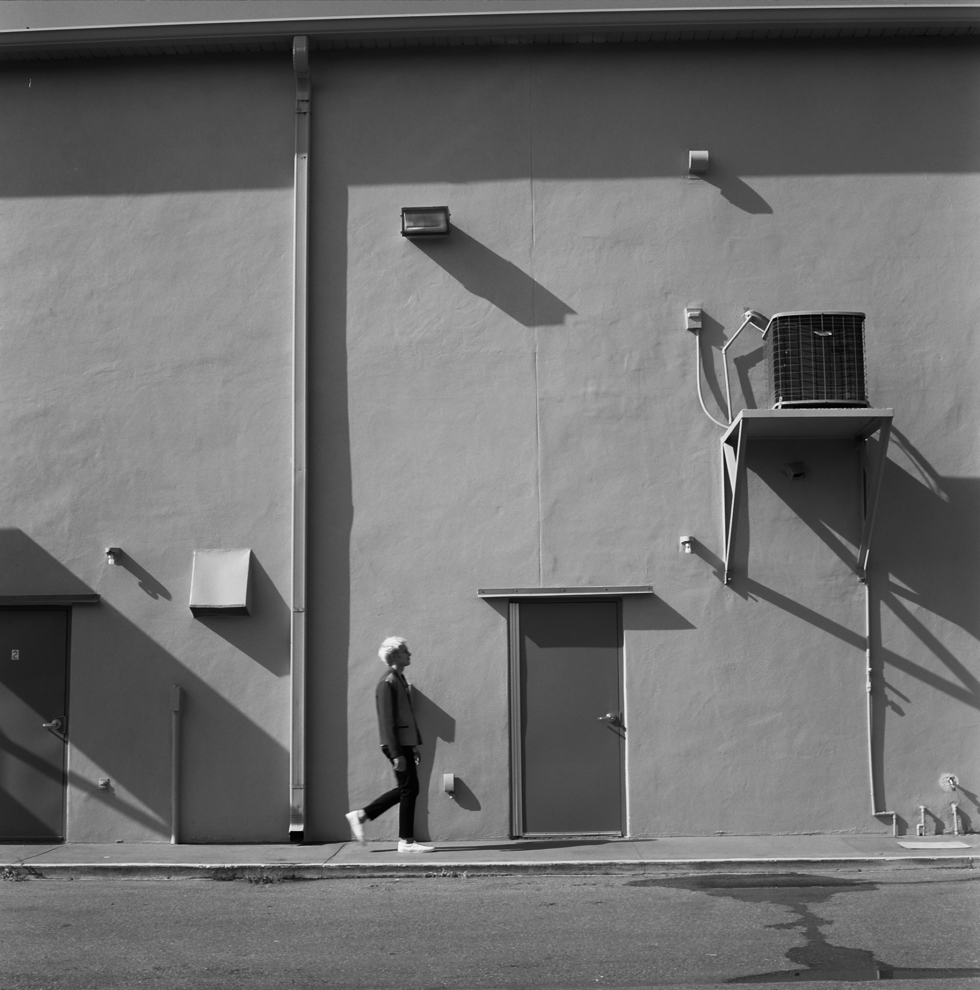 Person walking past a plain wall with a door, air conditioning unit, and shadows, in black and white.