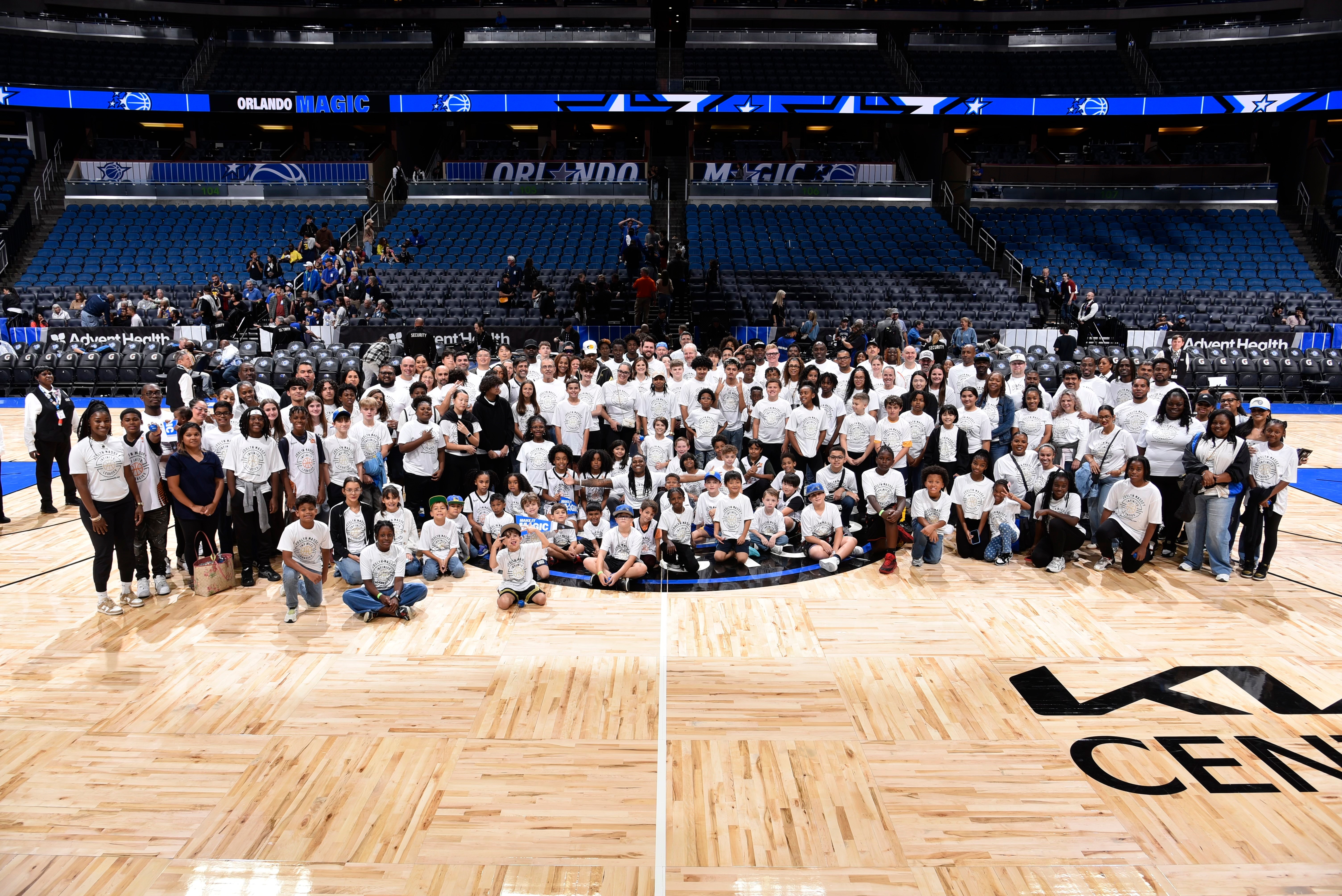 Large group of people gathered on basketball court inside an arena, with empty seats in the background.