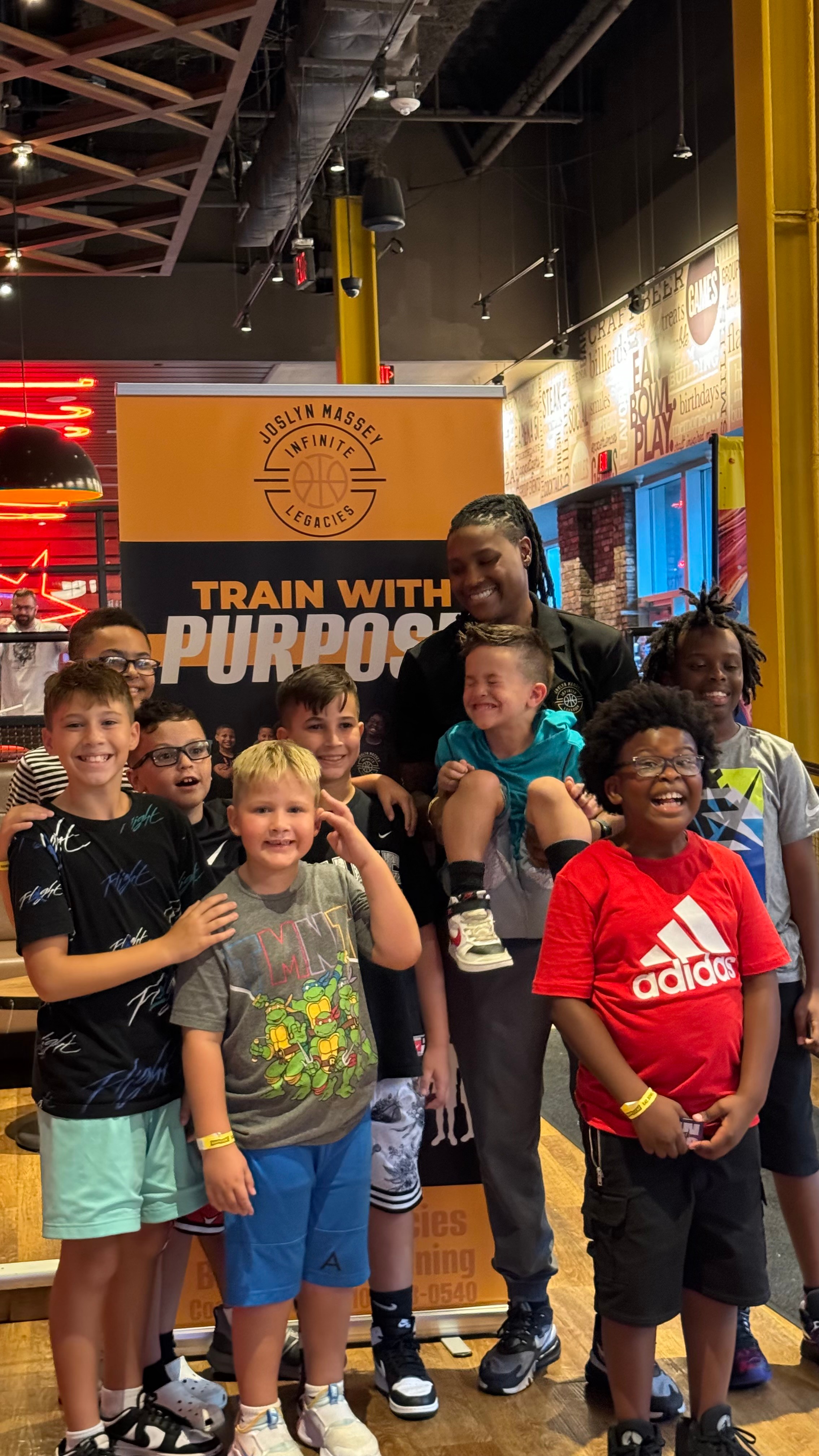 Group of children and a woman posing indoors with a banner behind them, some smiling and making gestures.
