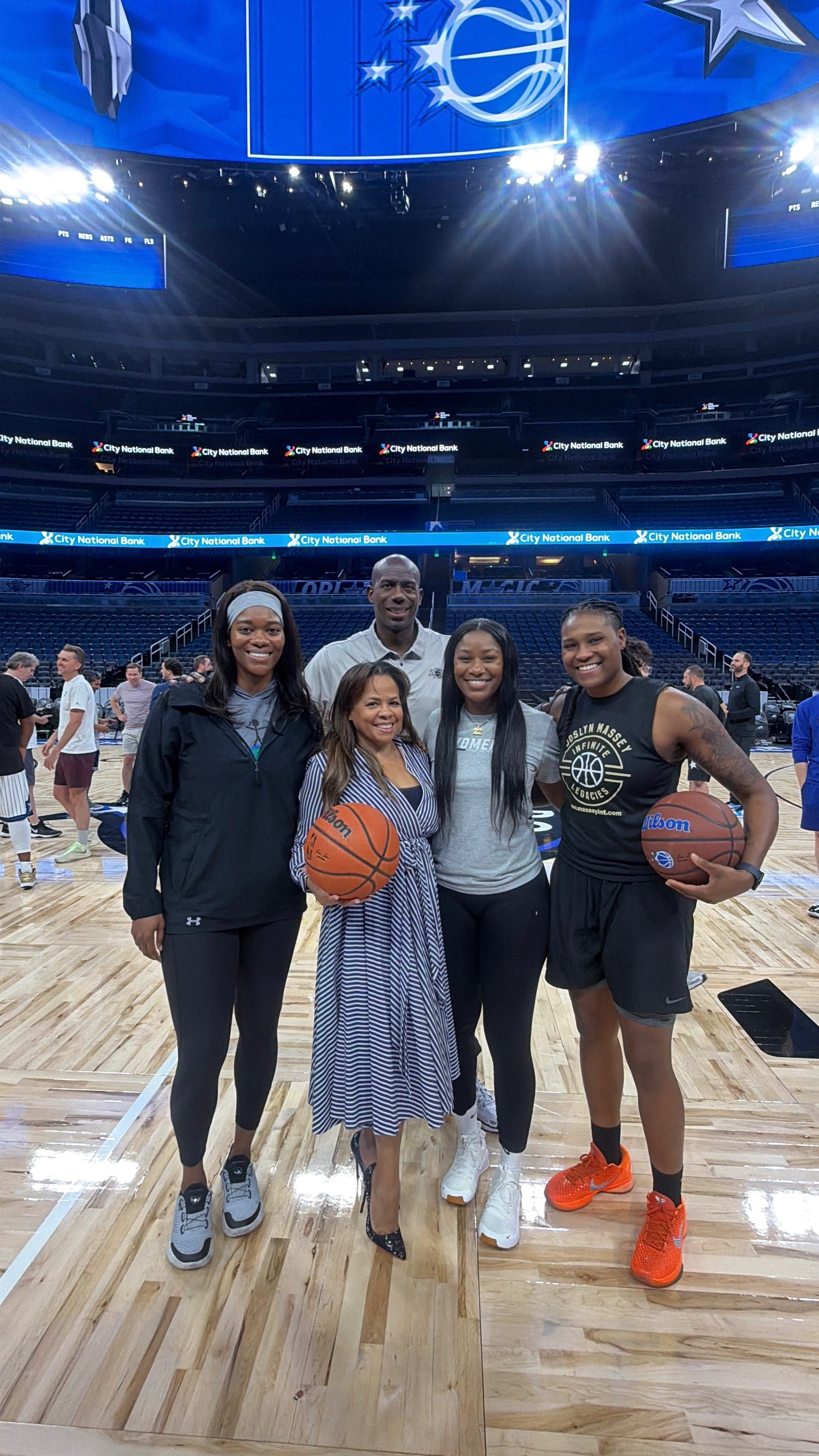 Five people standing on a basketball court inside a stadium, holding basketballs, smiling, with blue banners overhead.
