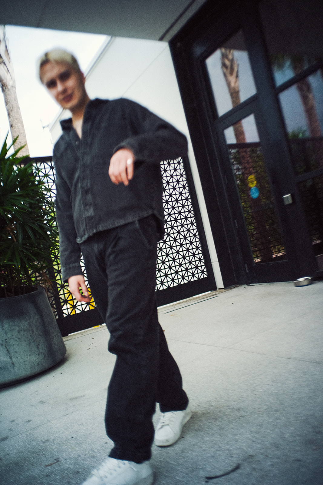 Person with short hair wearing dark clothing and white shoes standing outdoors near a building with a patterned gate.