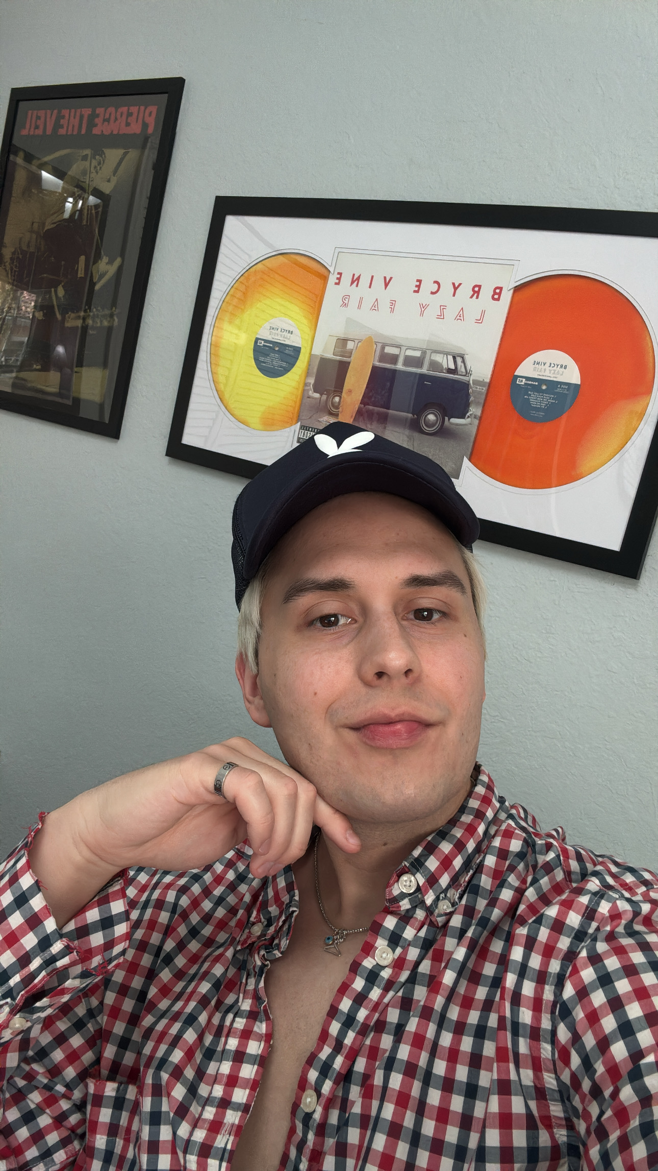 Young man wearing a checkered shirt and baseball cap, smiling, in front of framed artwork on a light-colored wall.