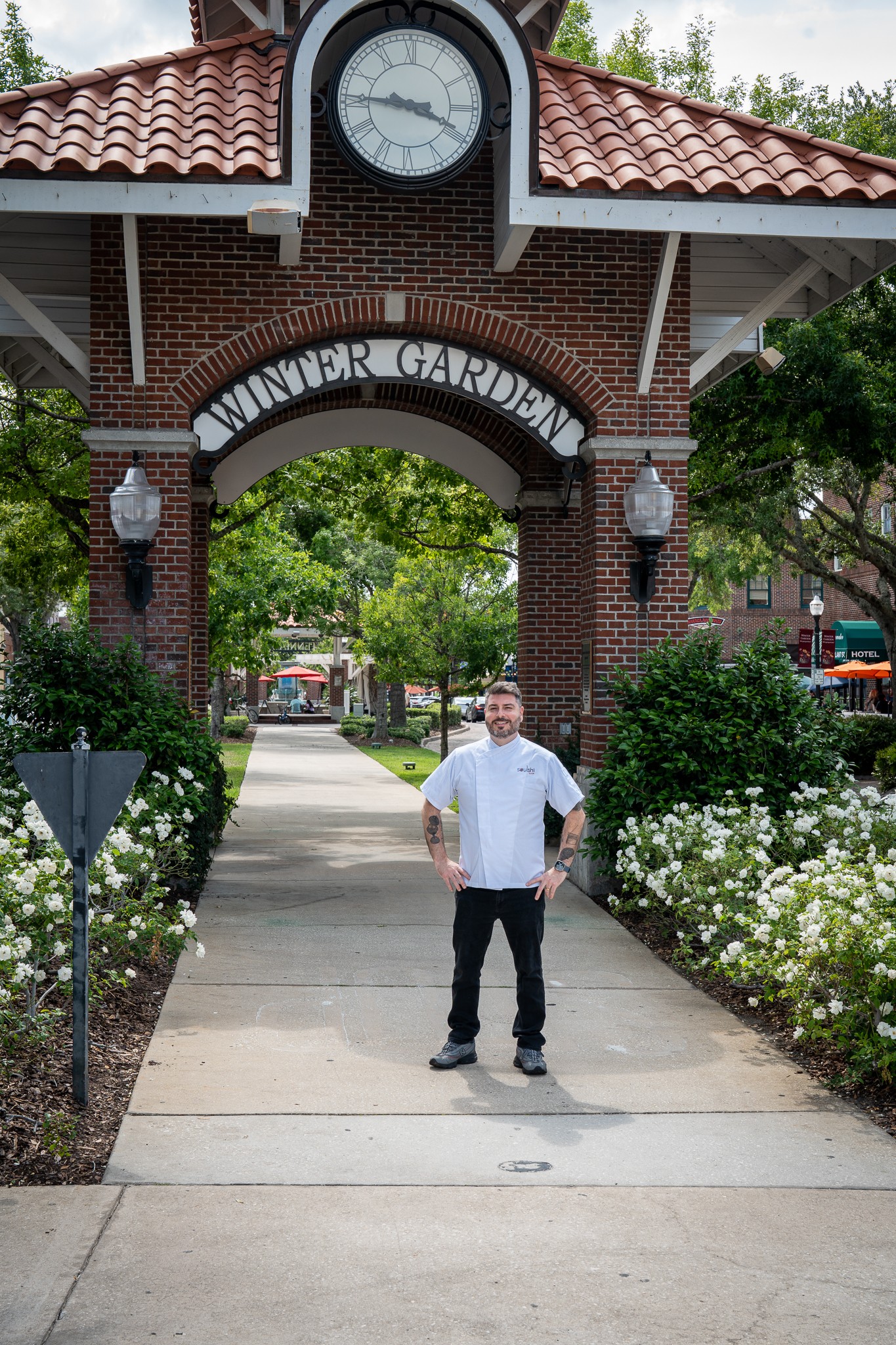 Person standing on sidewalk under brick archway with clock, greenery, and flowers, in a park entrance area.