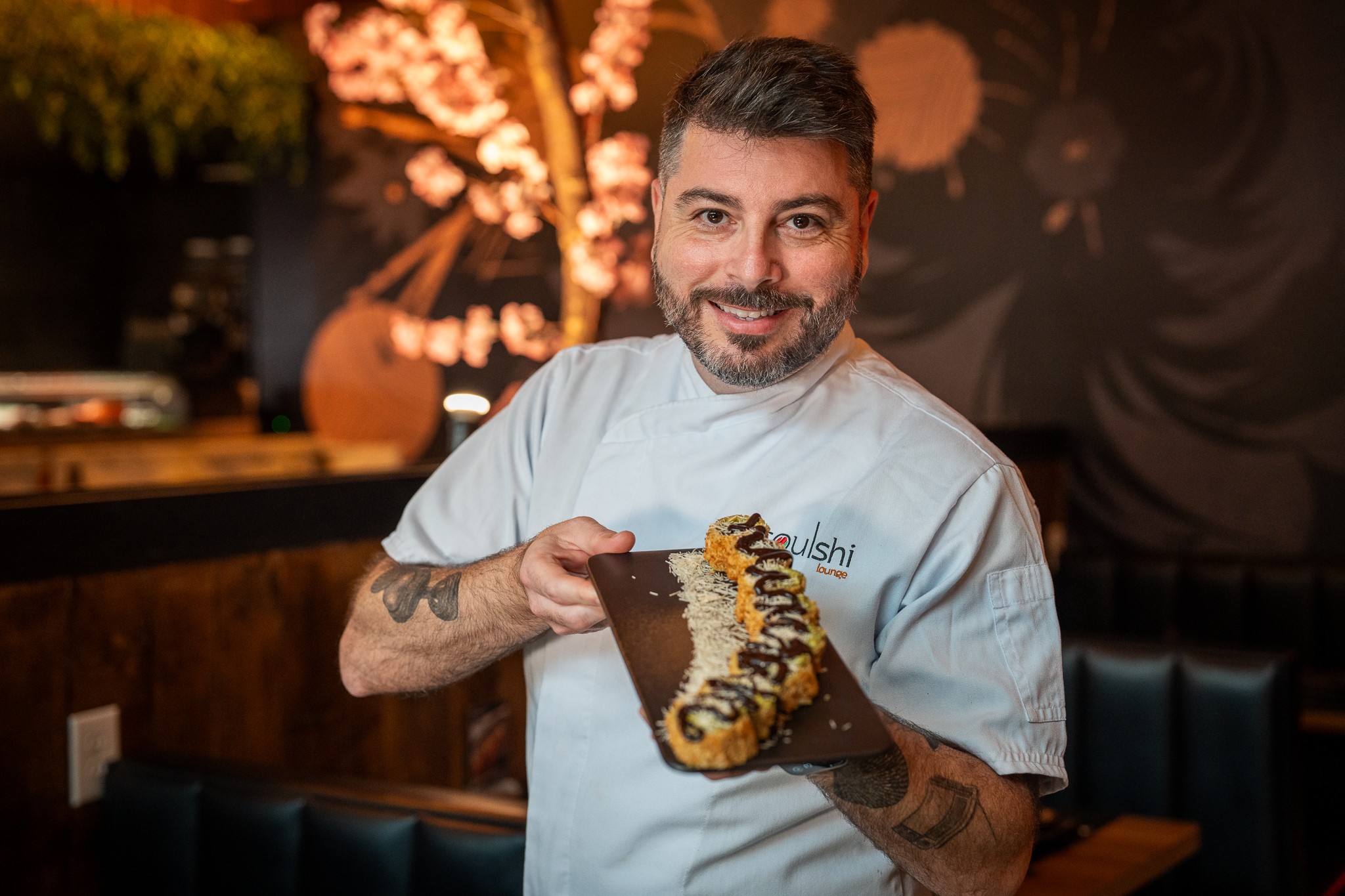 Man in chef's uniform holding a sushi roll with a smile, in a restaurant setting.
