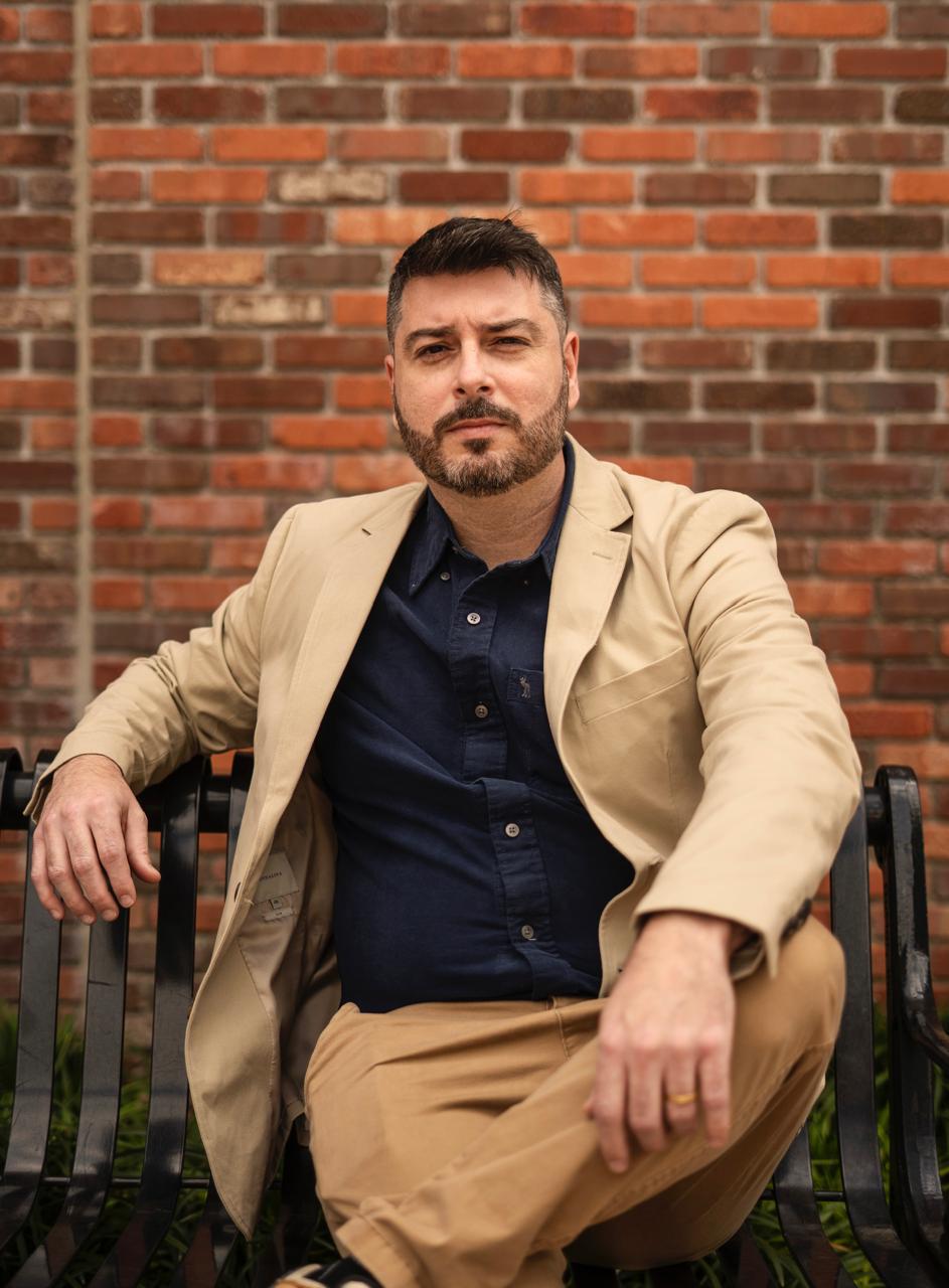 Man with dark hair and beard sitting on a black metal bench in front of a brick wall, wearing a beige blazer and navy shirt.