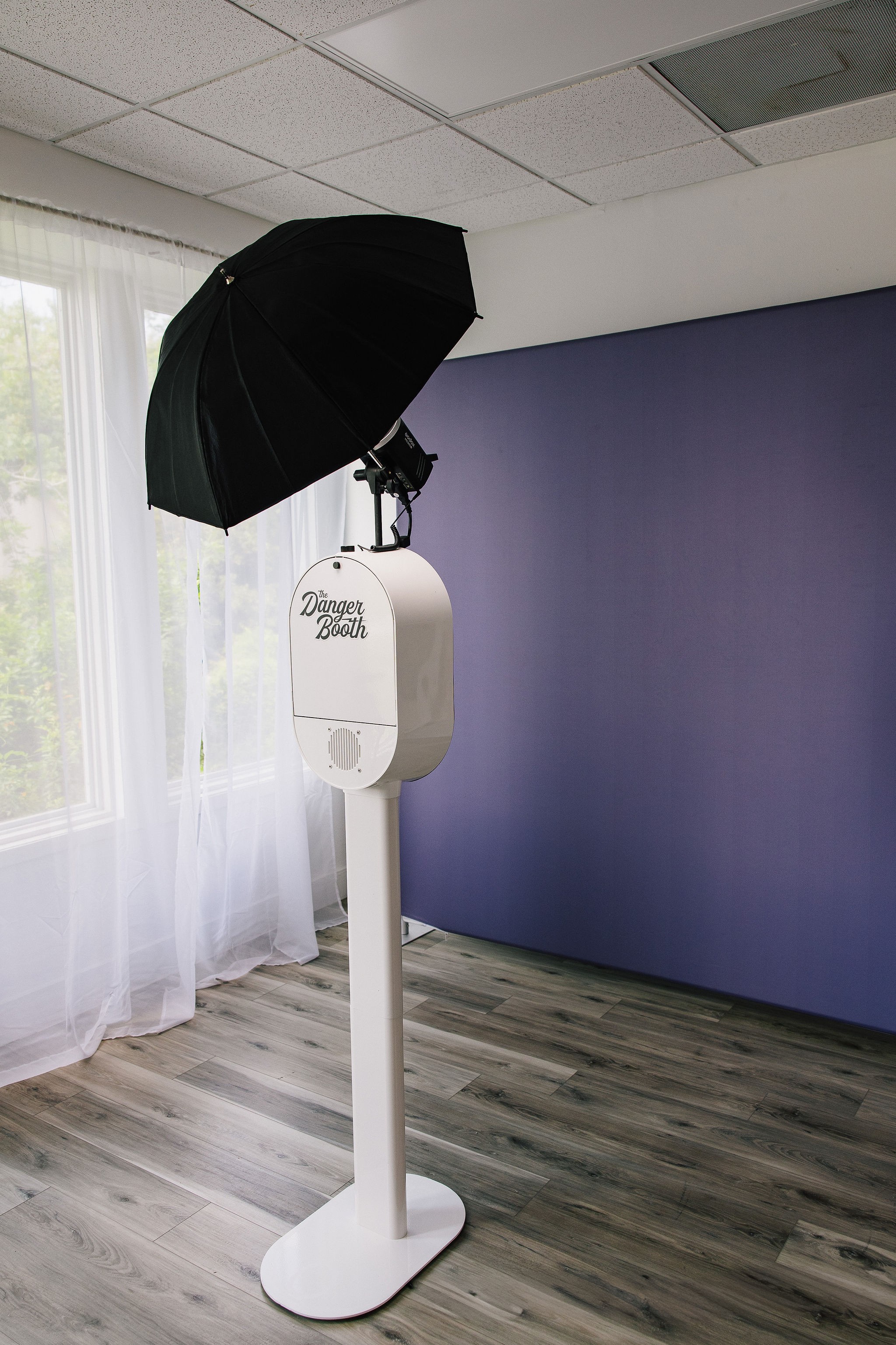 Photobooth setup with umbrella and backdrop in a room with wooden floor and window with curtains.