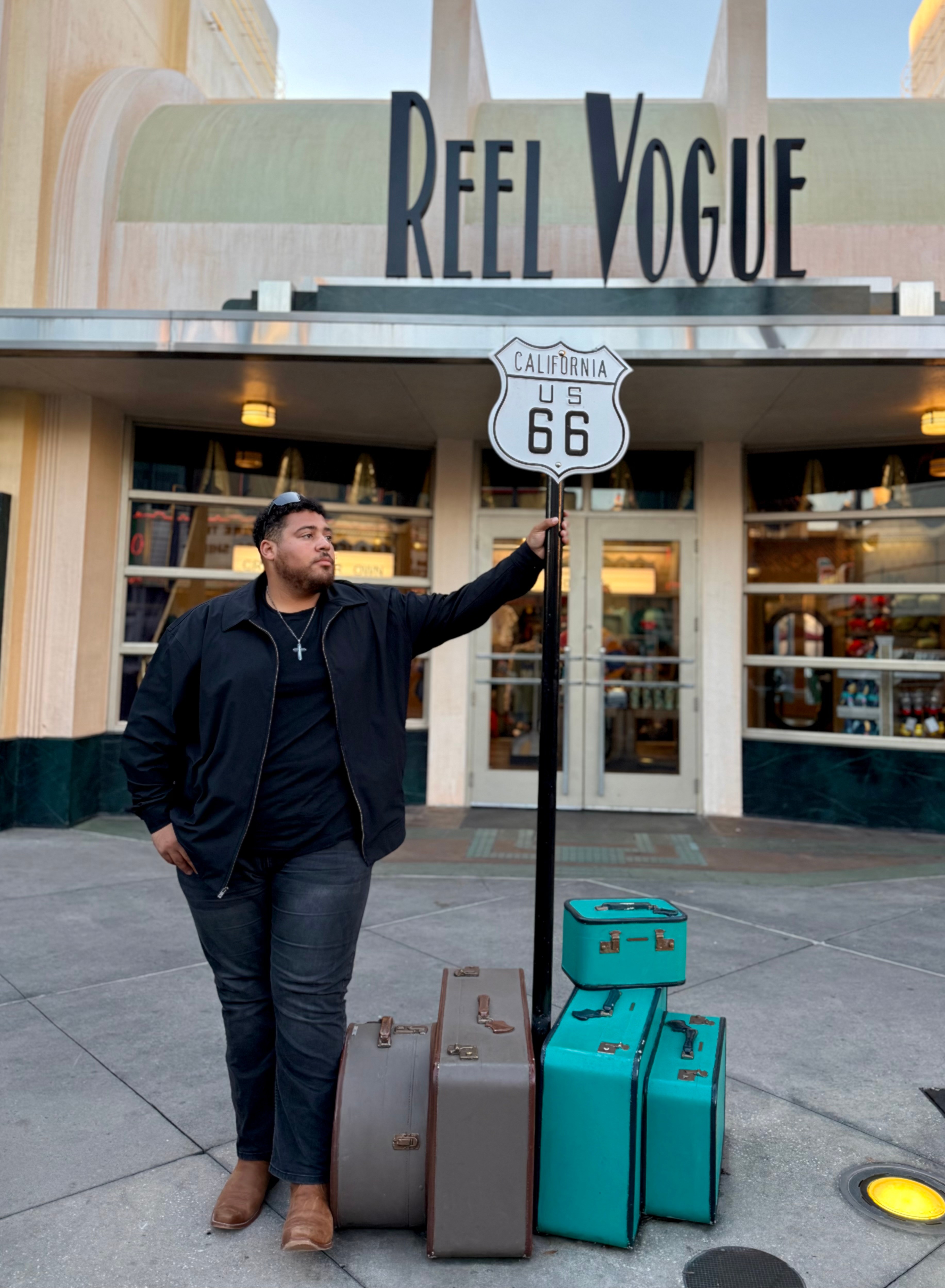 Man standing outside a store with suitcases, holding a Route 66 sign, in front of Reel Vogue shop, daytime.