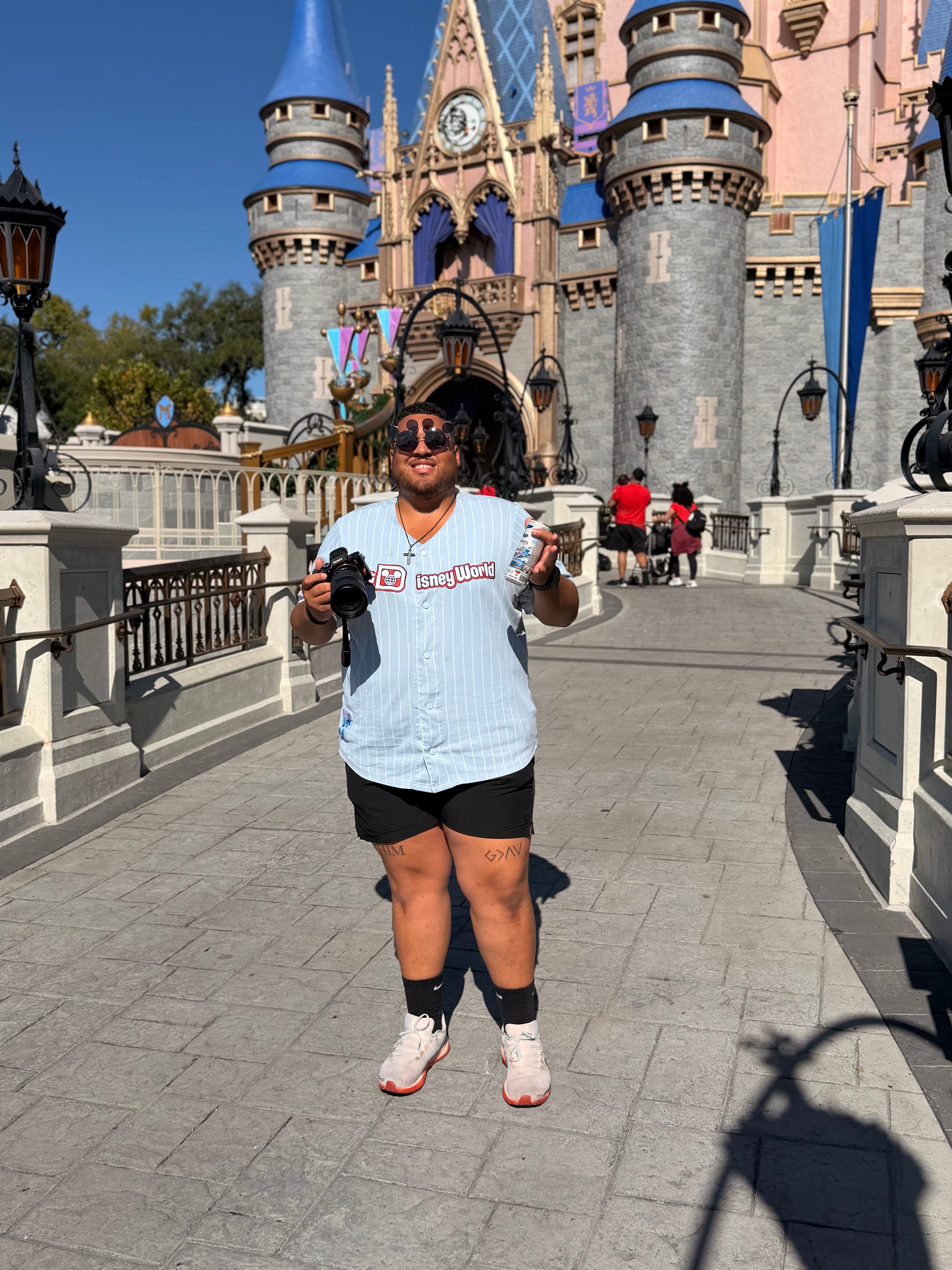 Person standing in front of a castle at a theme park, holding a drink and a snack, smiling, sunny day.