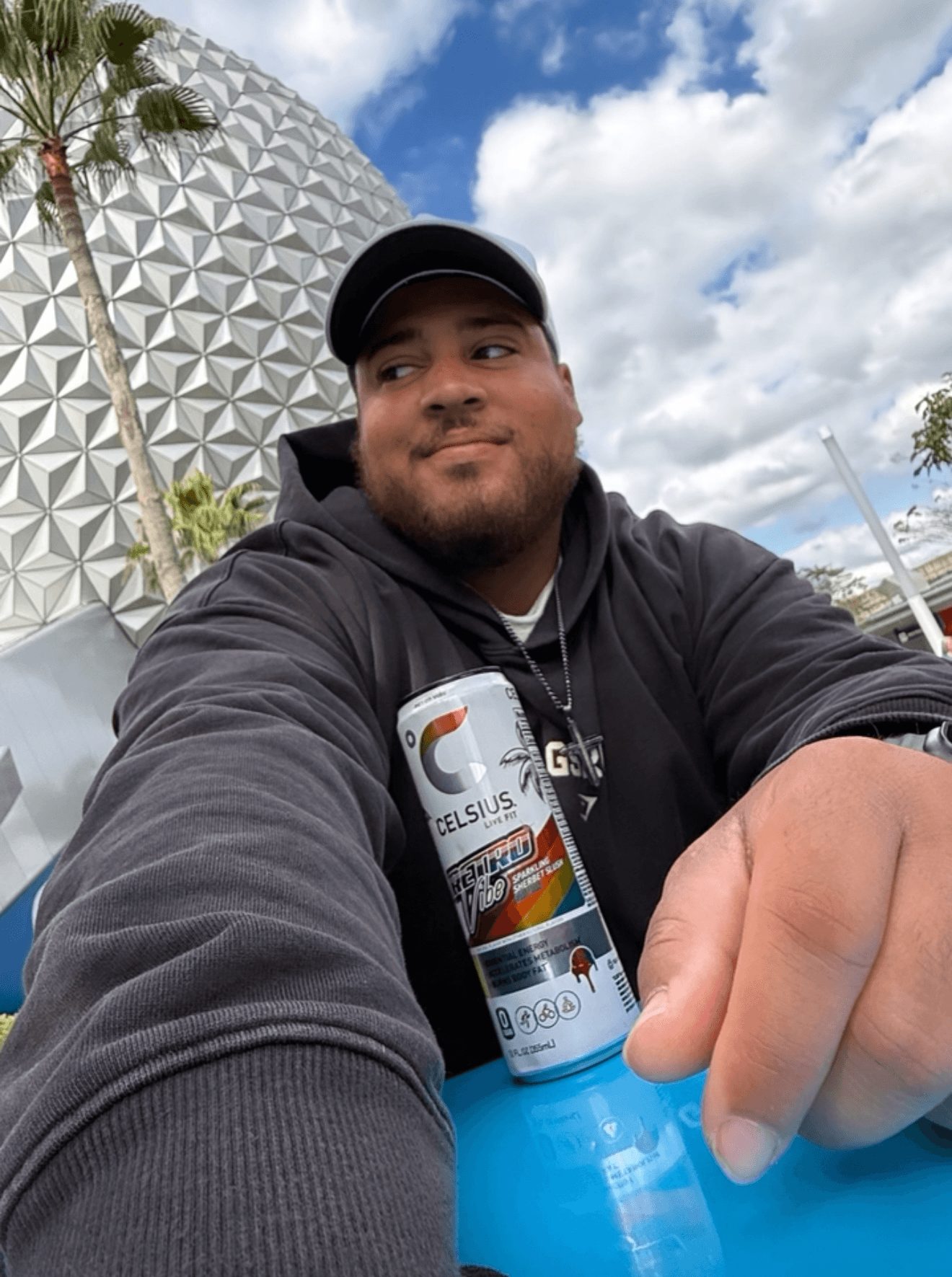Young man in black hoodie and cap outdoors, holding a can of Celsius energy drink, with a geodesic dome and cloudy sky in background.