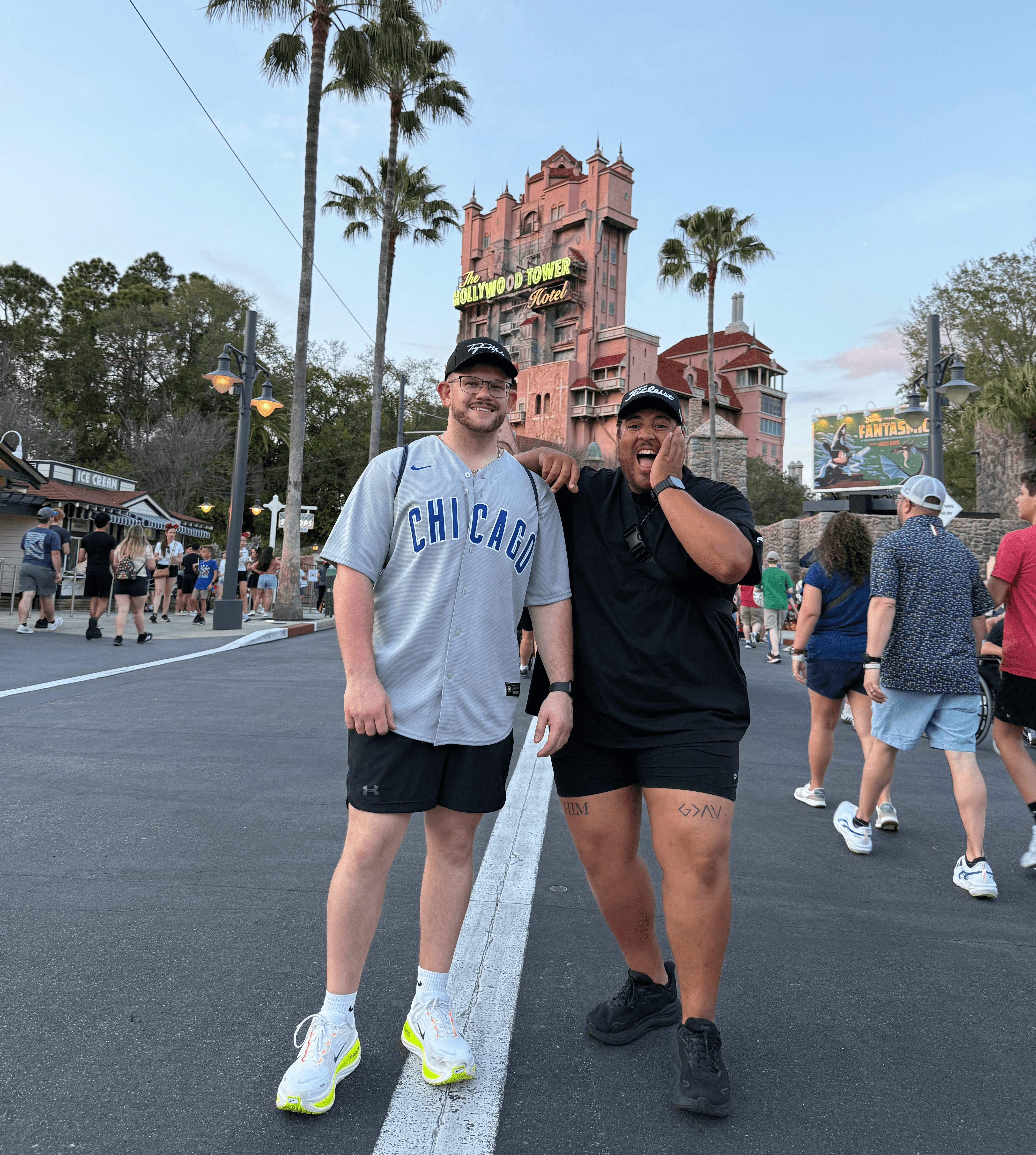 Two people smiling and posing in front of a castle at a theme park, surrounded by other visitors, palm trees, and a clear sky.