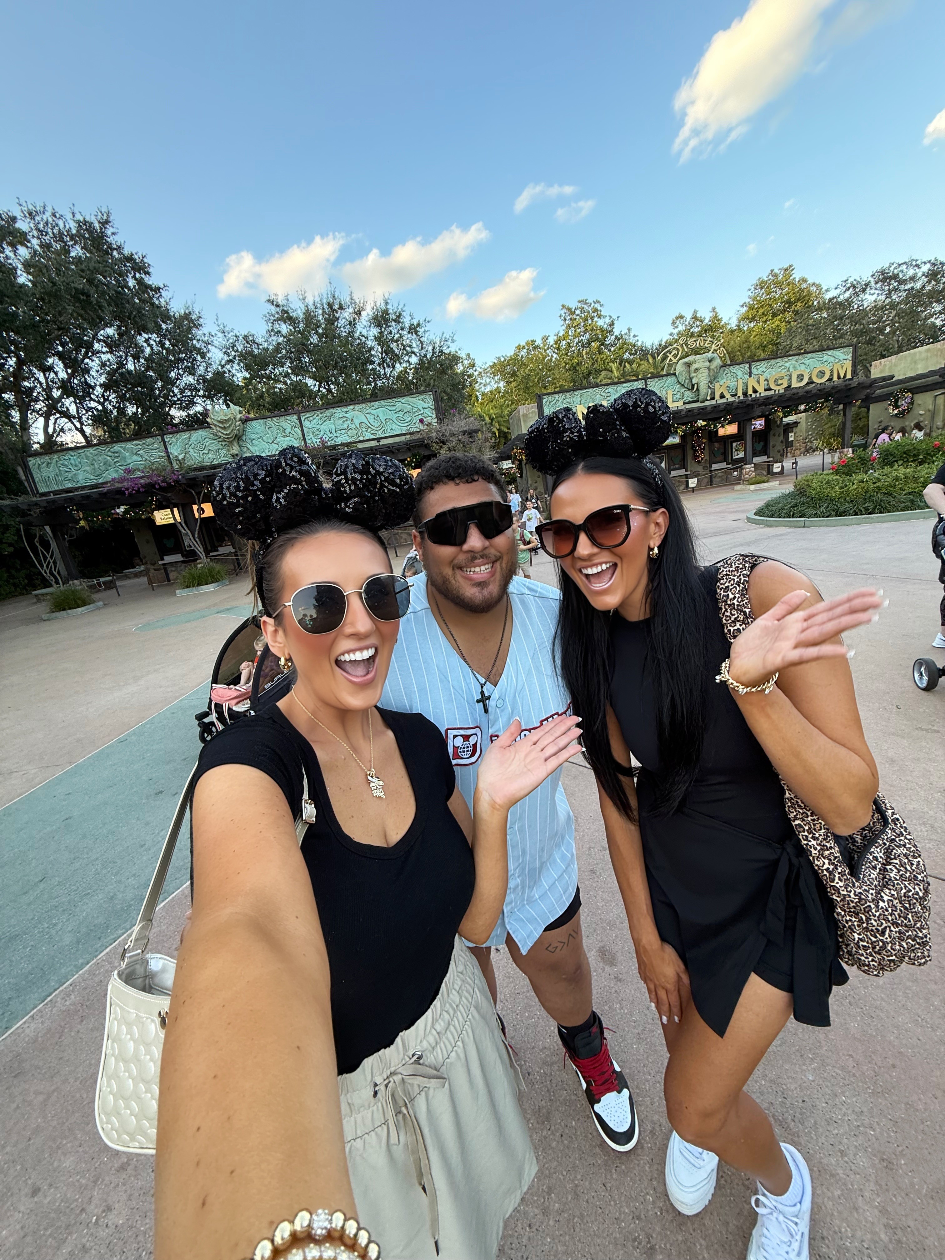 Three people smiling and posing outdoors at a park or zoo, with trees and a building in the background.