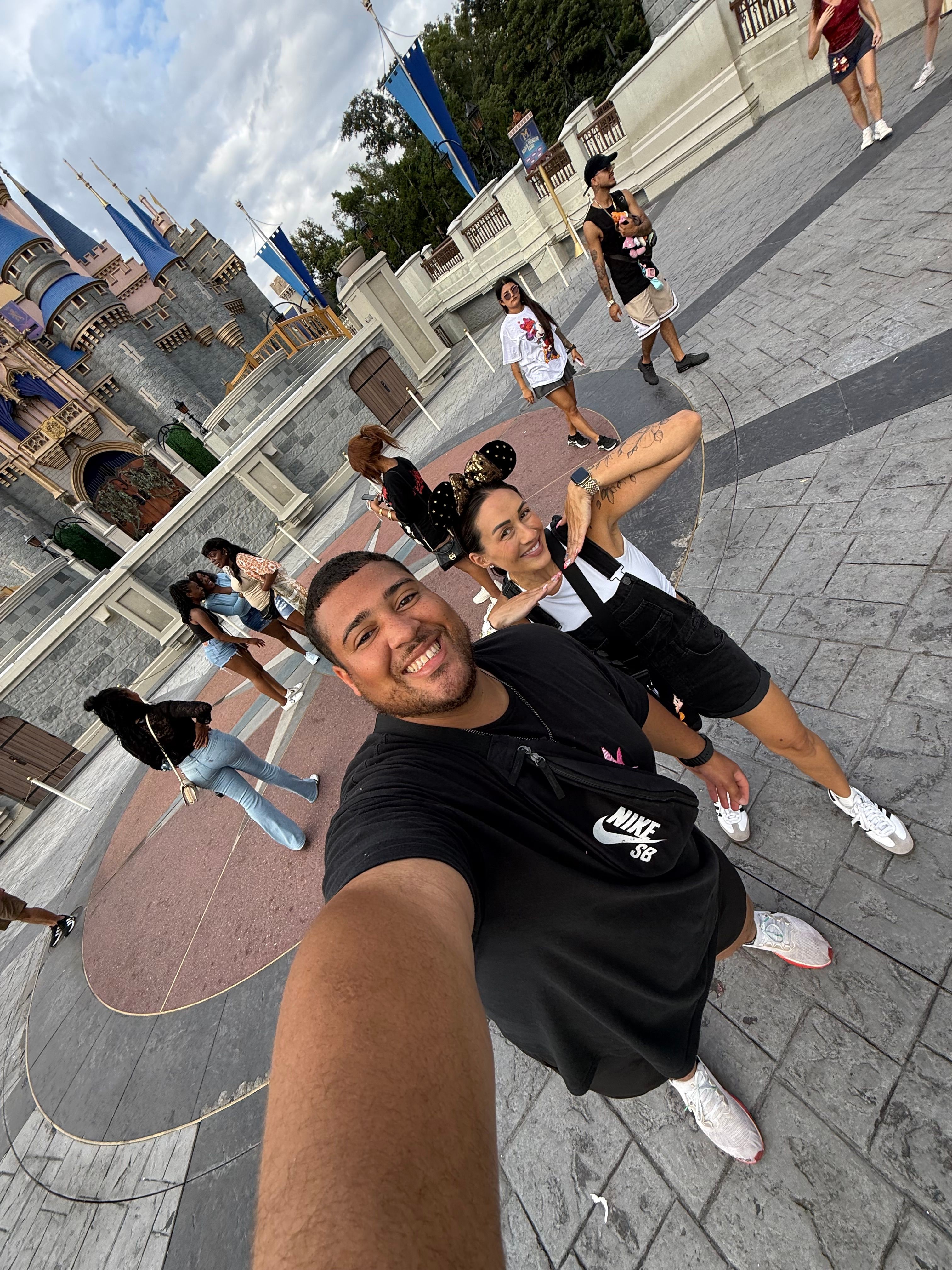 Person taking a selfie on a city sidewalk with buildings and people in the background.