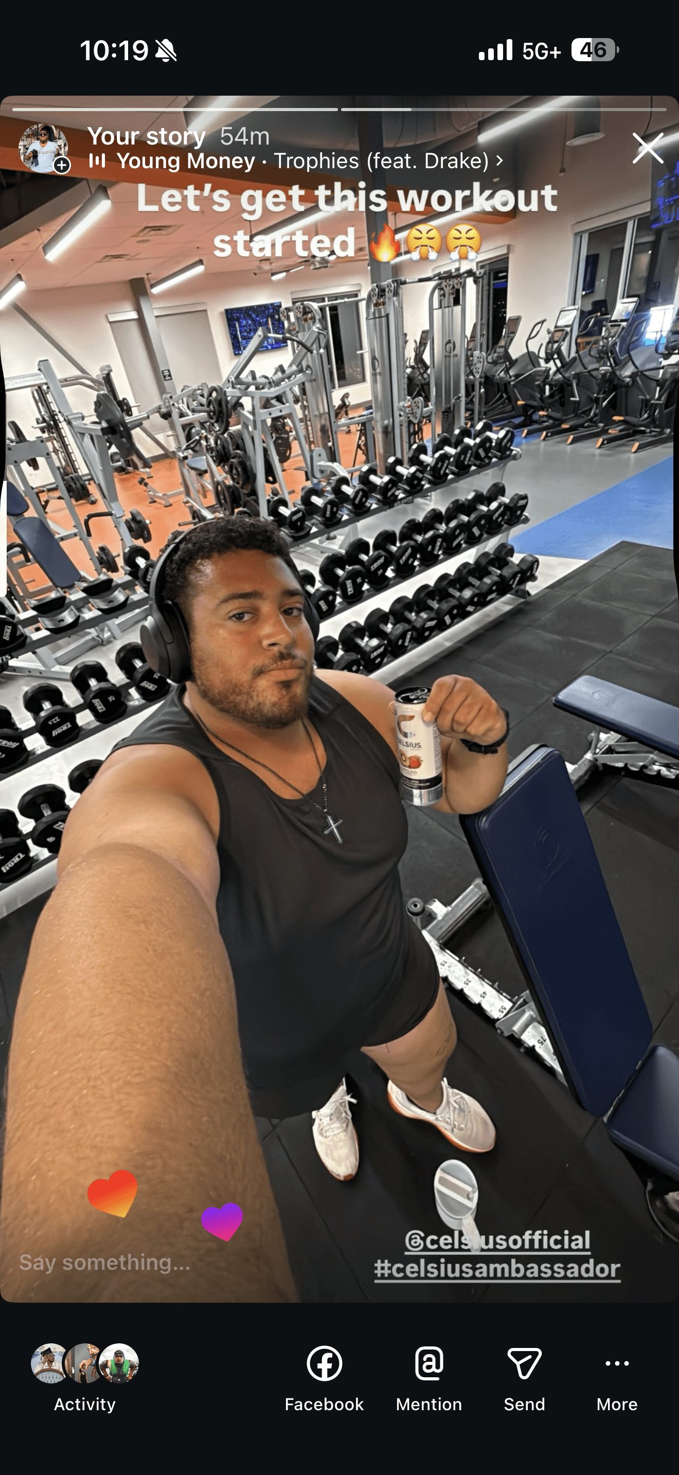 Person taking selfie in gym with weights and exercise equipment in background, holding a drink, wearing black workout clothes.