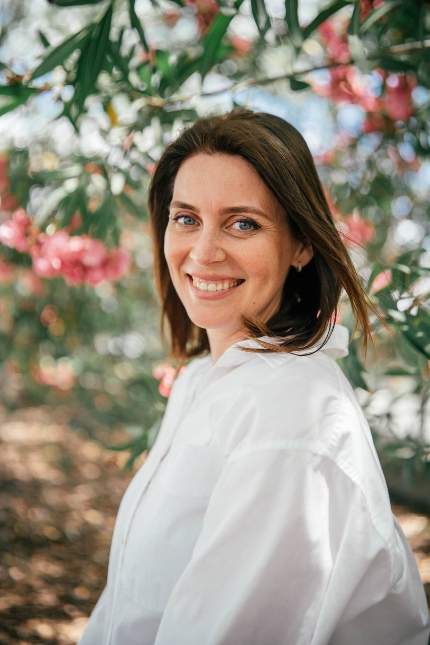 Smiling woman with shoulder-length dark hair outdoors among pink flowers and green foliage.