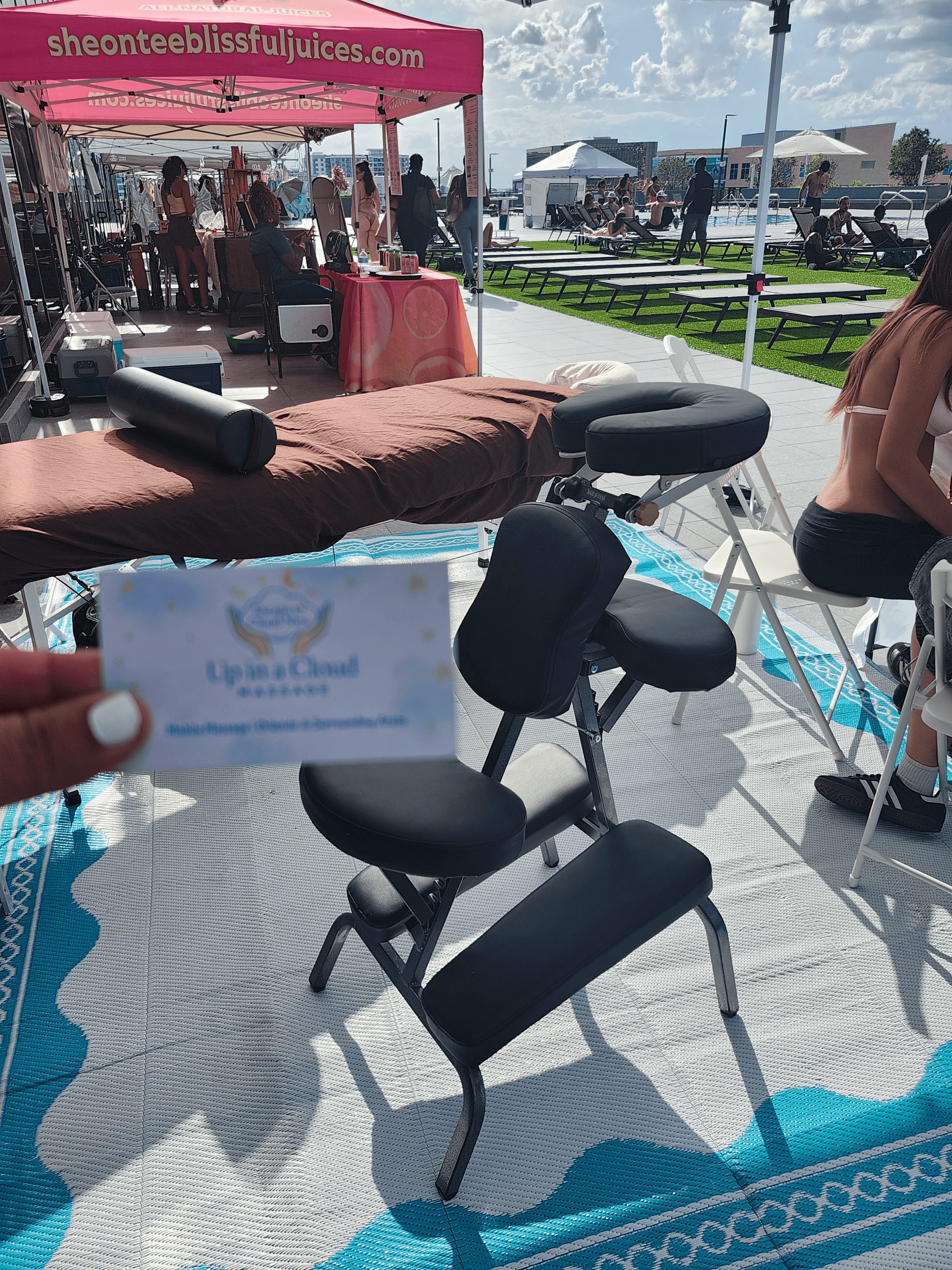 Massage chair with headrest and armrest outdoors, with tents and people in the background on a sunny day.