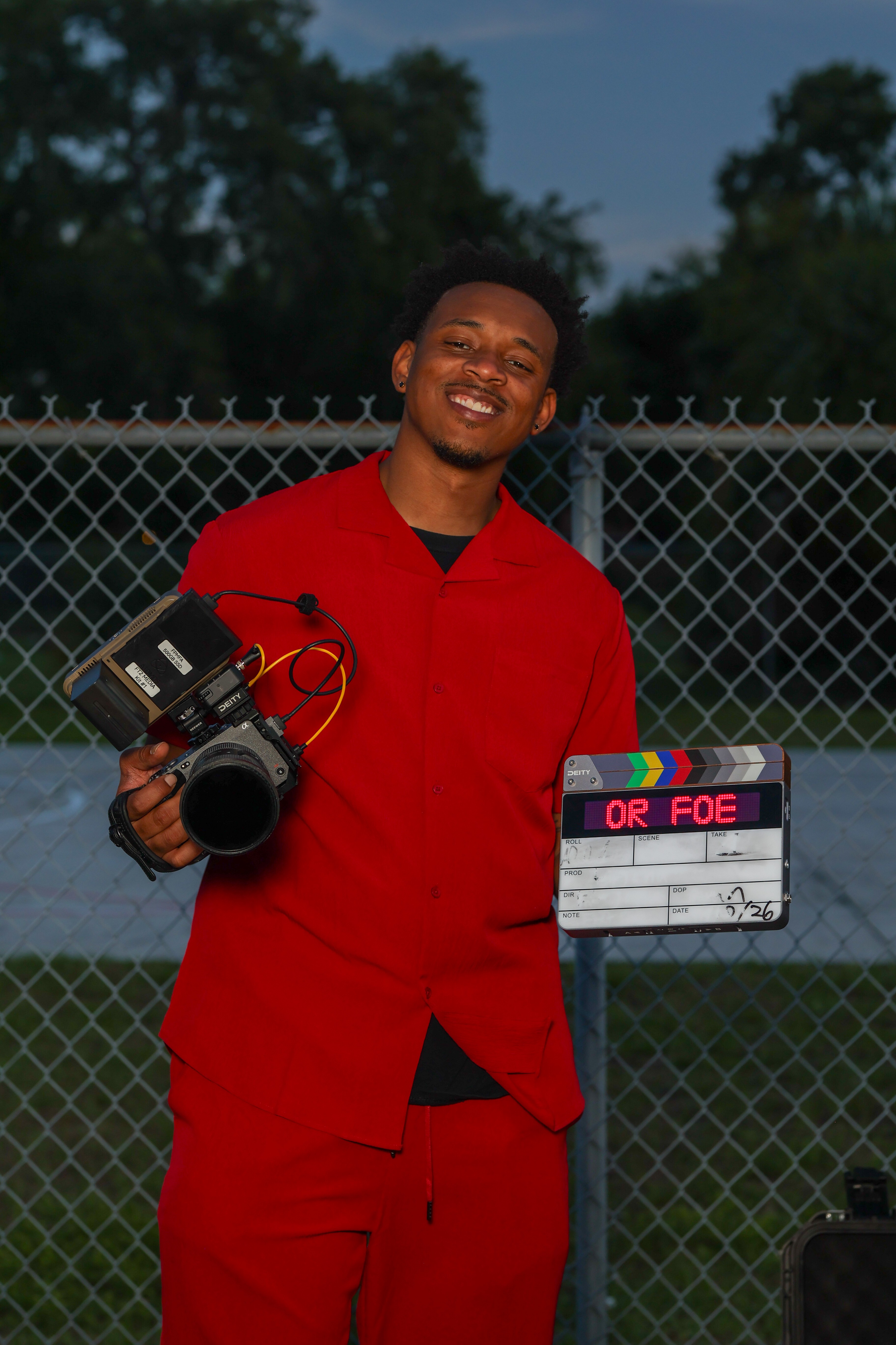 Young man in red sports uniform holding a camera and a clapperboard outdoors, smiling, with trees and a fence in background.