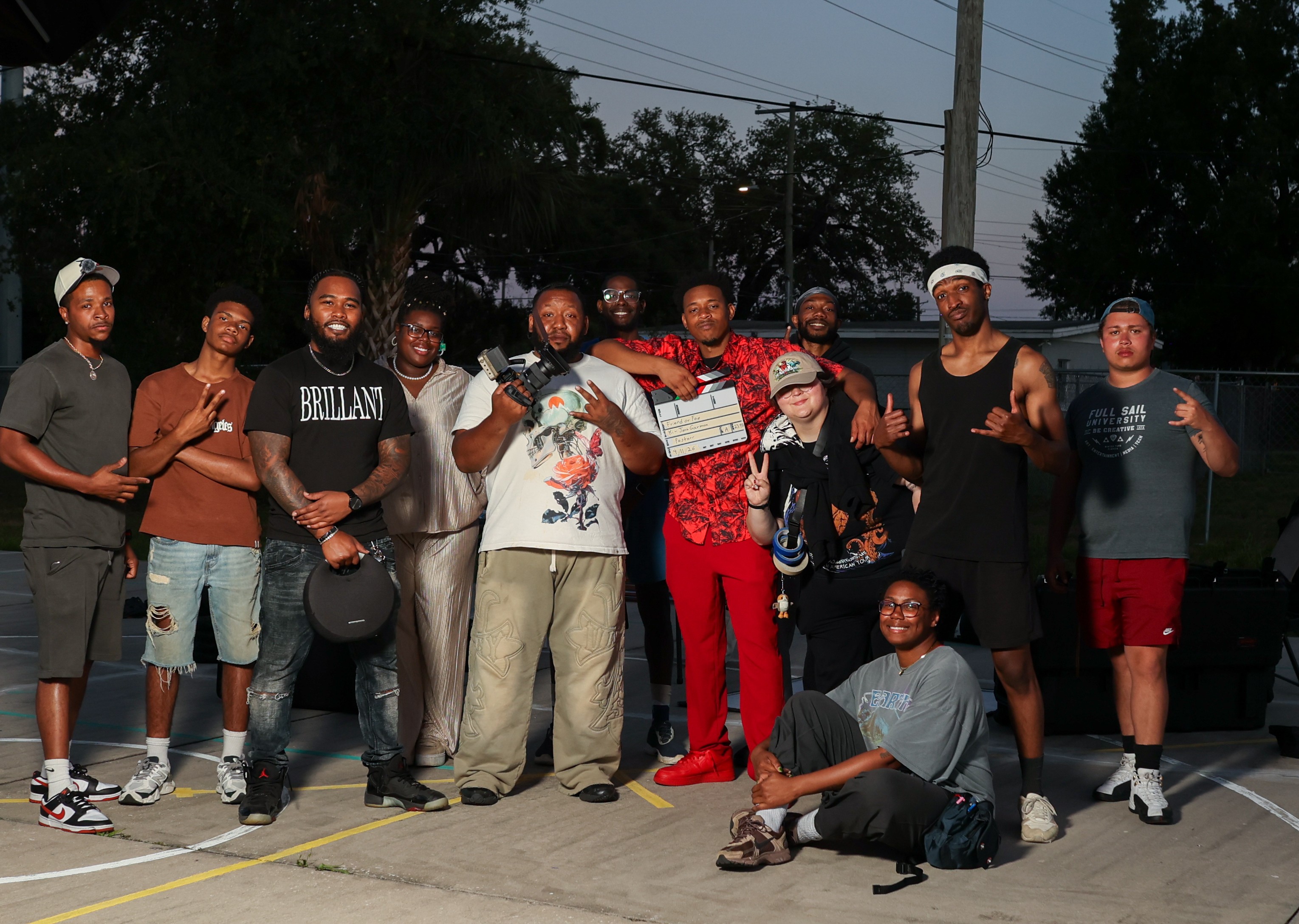 Group of 12 people standing outdoors on pavement, some smiling, some making gestures, trees and power lines in background.