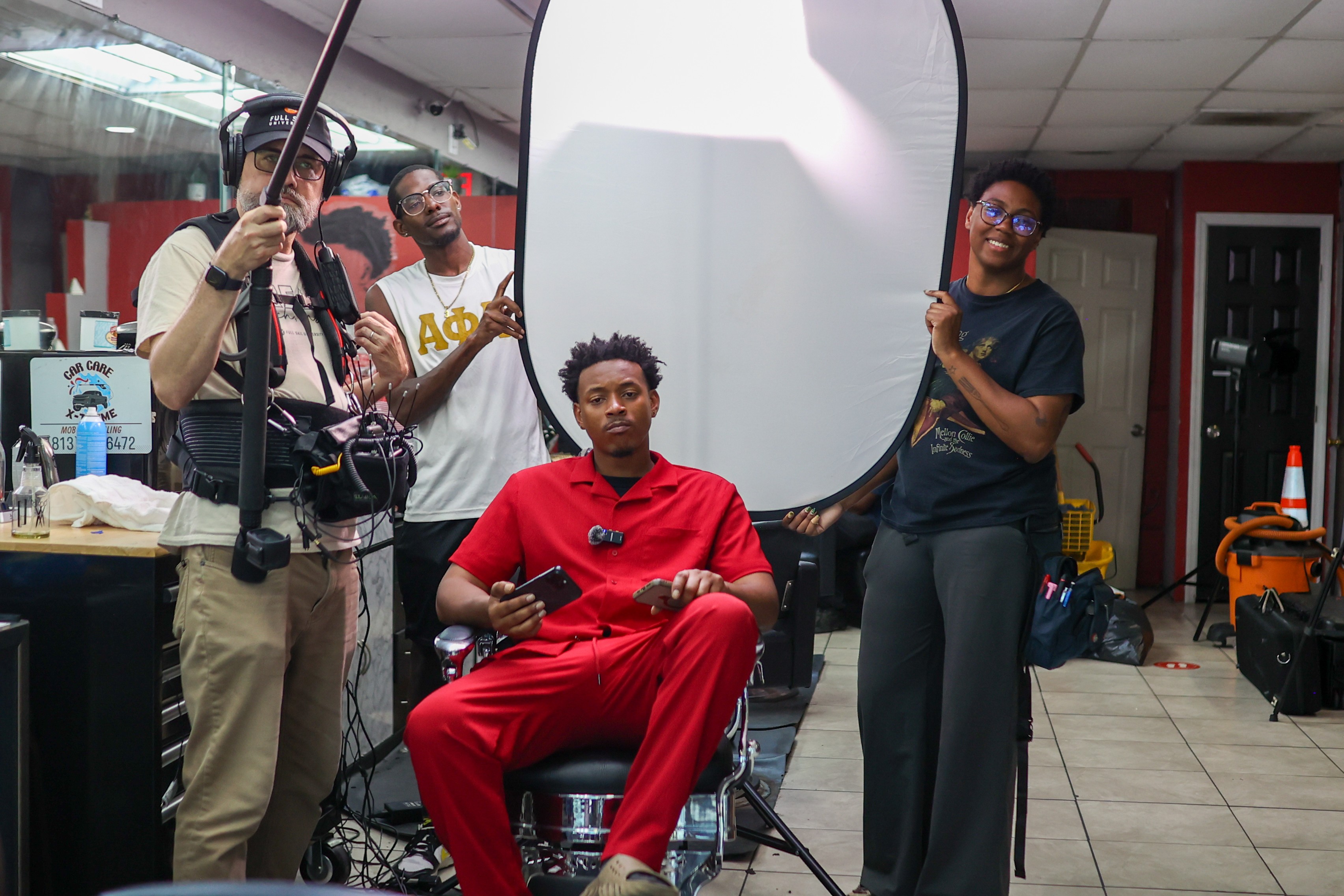 Person in red clothing seated in a chair with crew members setting up a photo shoot, large reflector, and equipment visible.