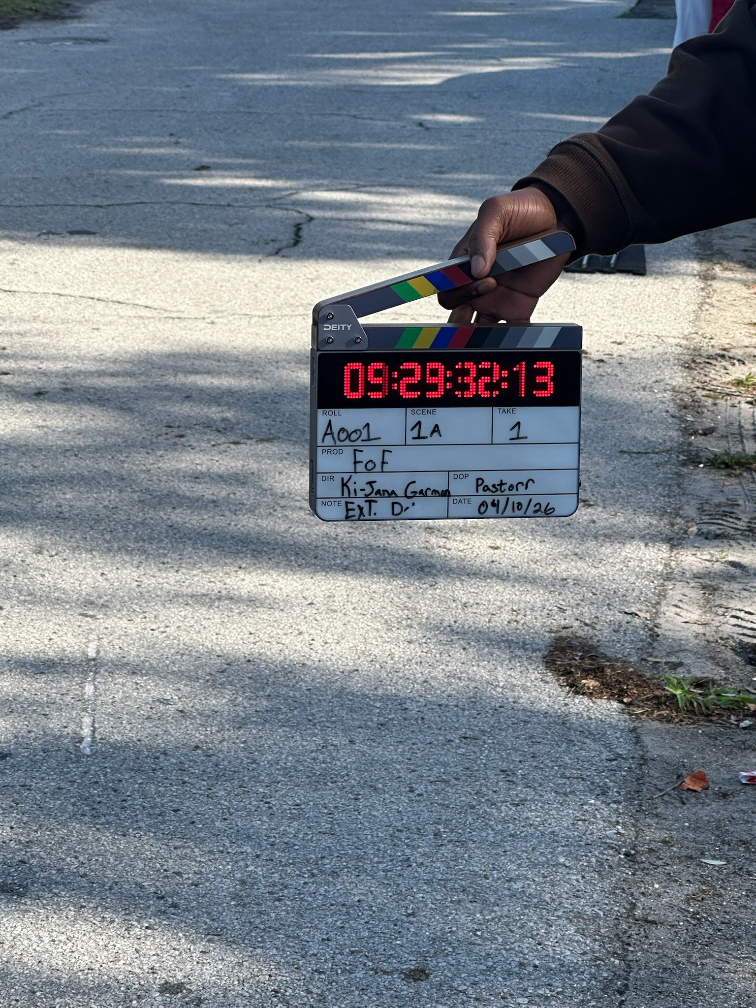 Person holding a clapperboard with a digital timer, on a street with shadows and cracks, hand wearing a dark sleeve.