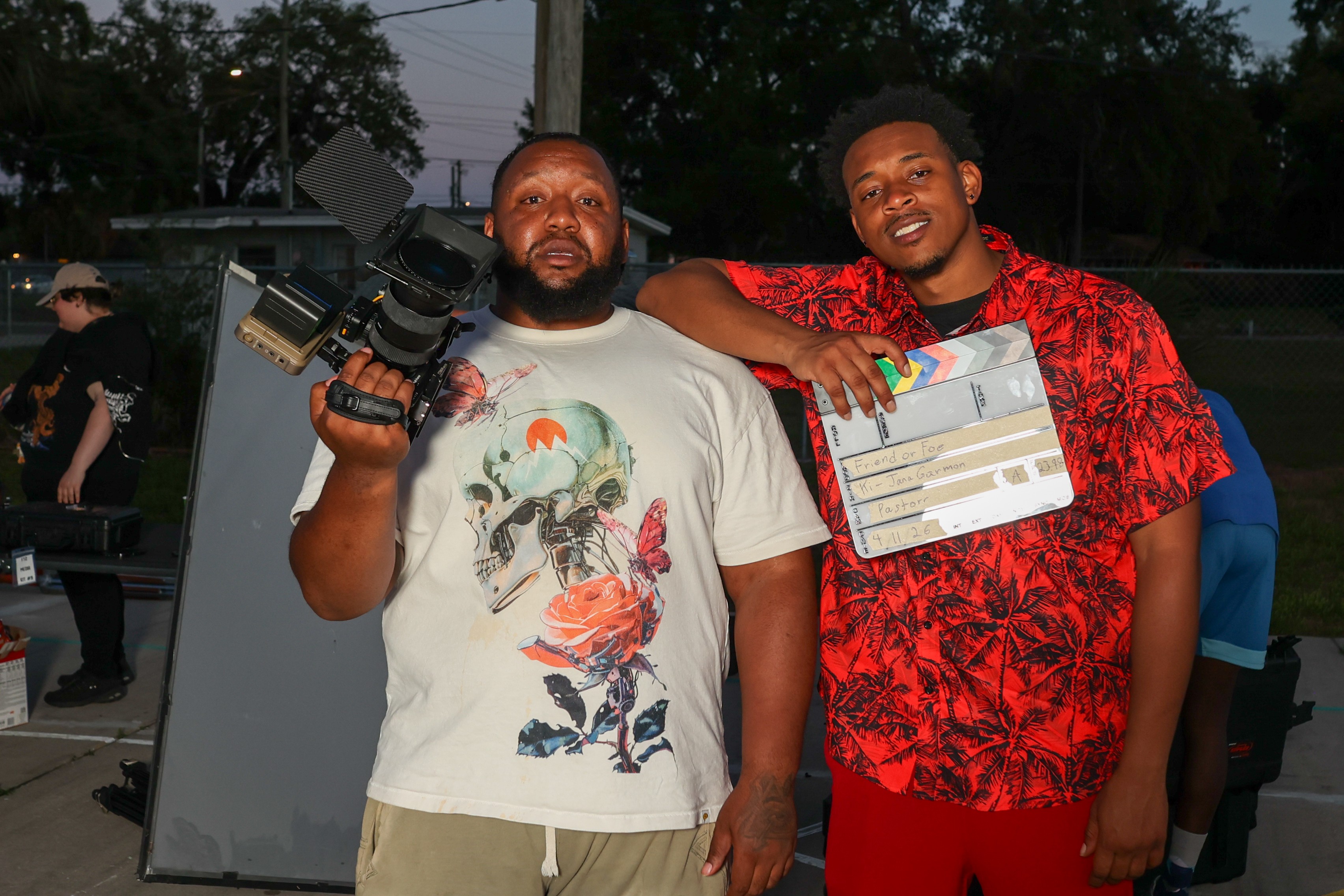 Two men standing outdoors, one holding a camera, the other holding a clapperboard, with trees and people in the background.