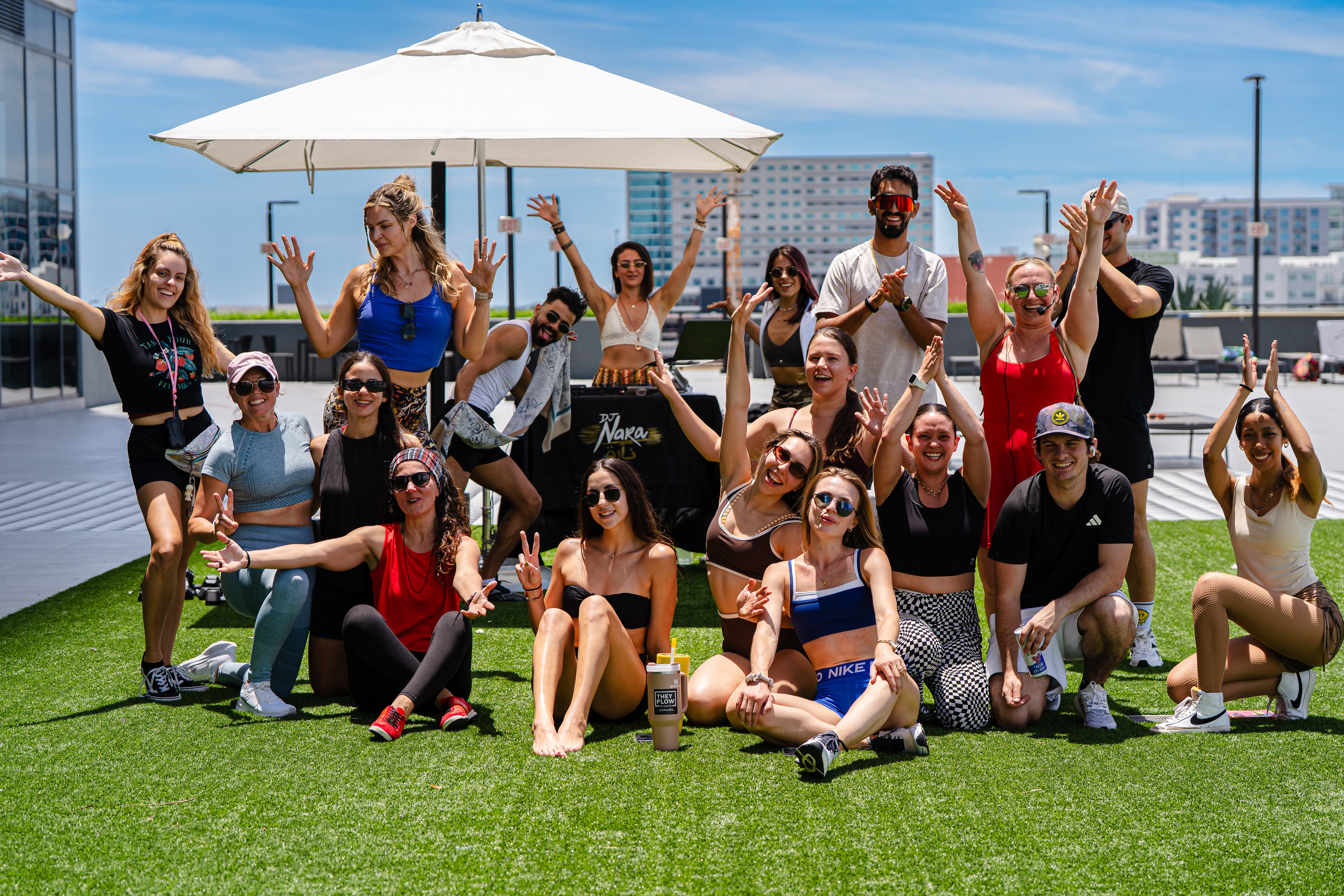 Group of people posing on a rooftop with a city view, some standing and some sitting on grass, under a large umbrella.