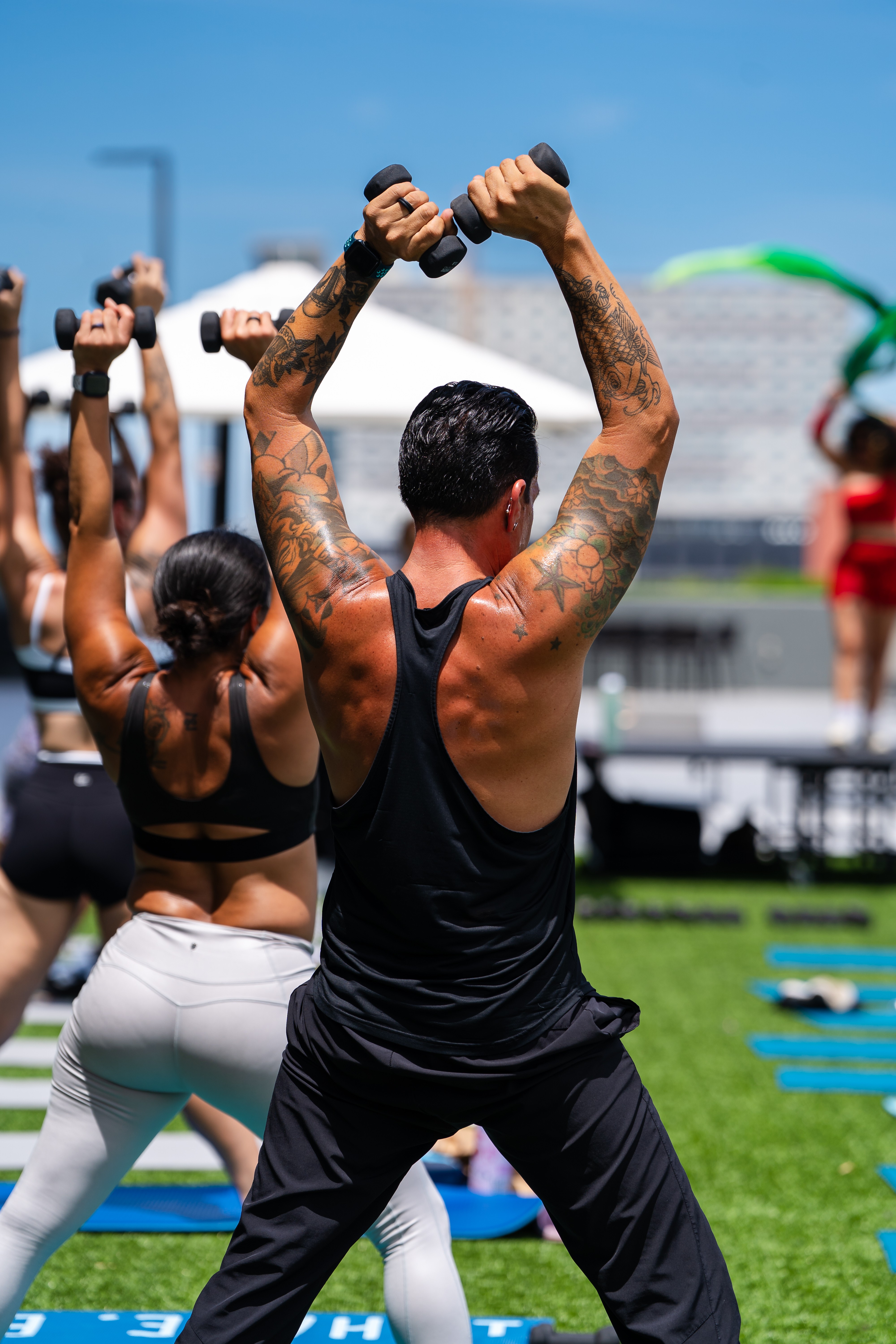 People exercising outdoors, raising their arms with dumbbells, on a sunny day with a stadium in the background.