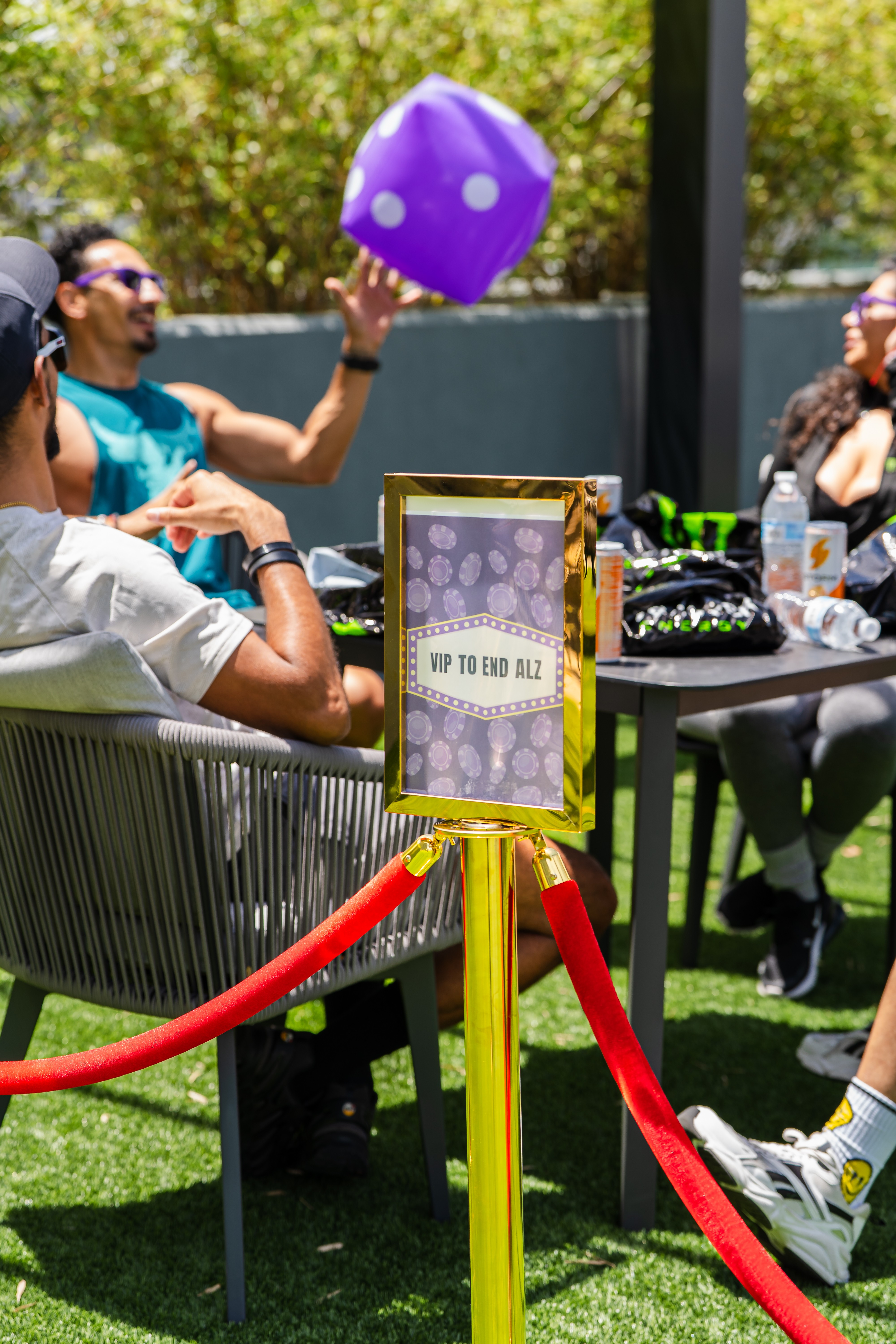 People sitting outdoors at a table with a large purple die, surrounded by trees and a red rope barrier.