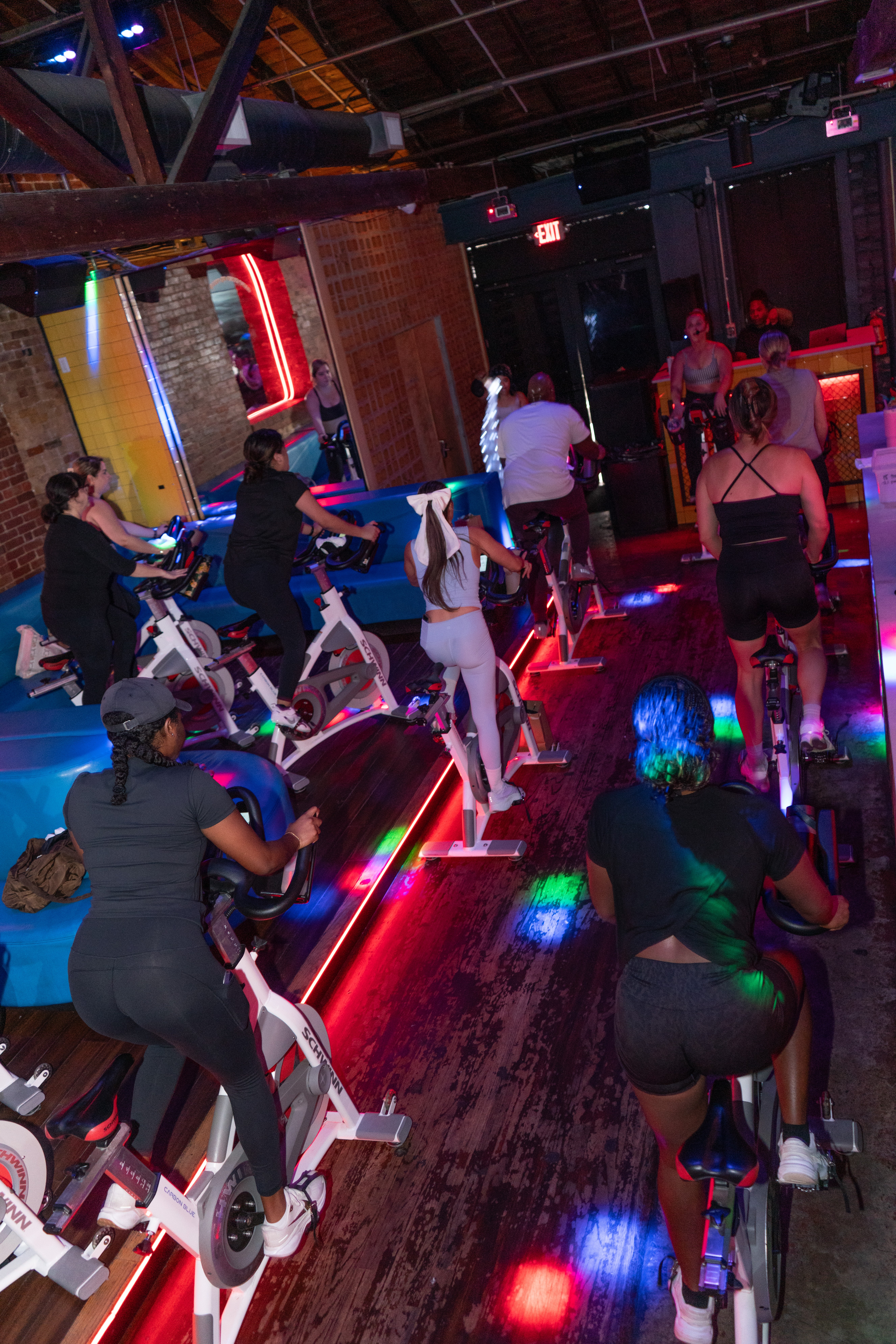 People riding stationary bikes in a dimly lit room with colorful lights and a brick wall background.