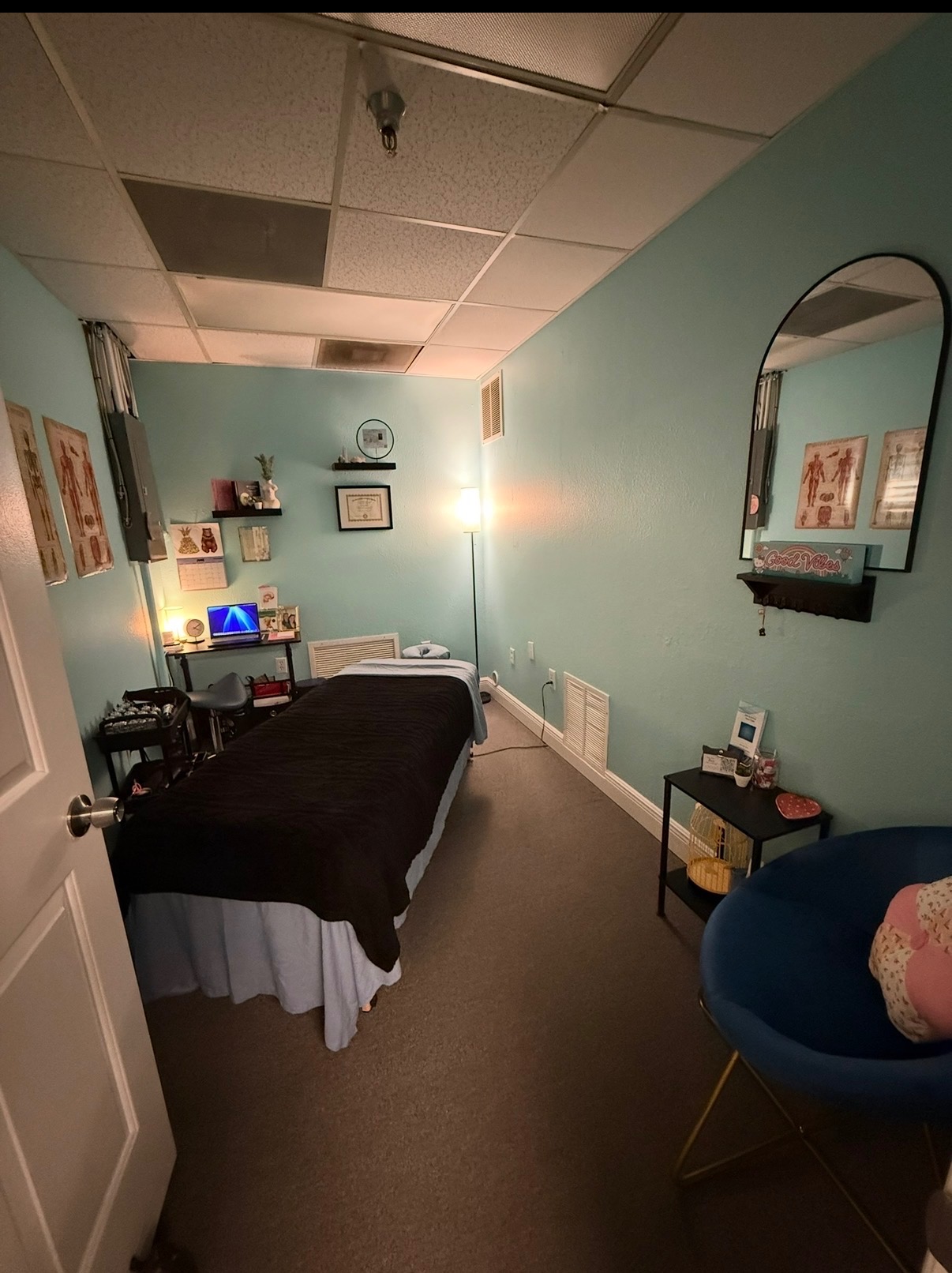 A massage room with a bed, mirror, and seating area, illuminated by a lamp, with light blue walls and ceiling tiles.