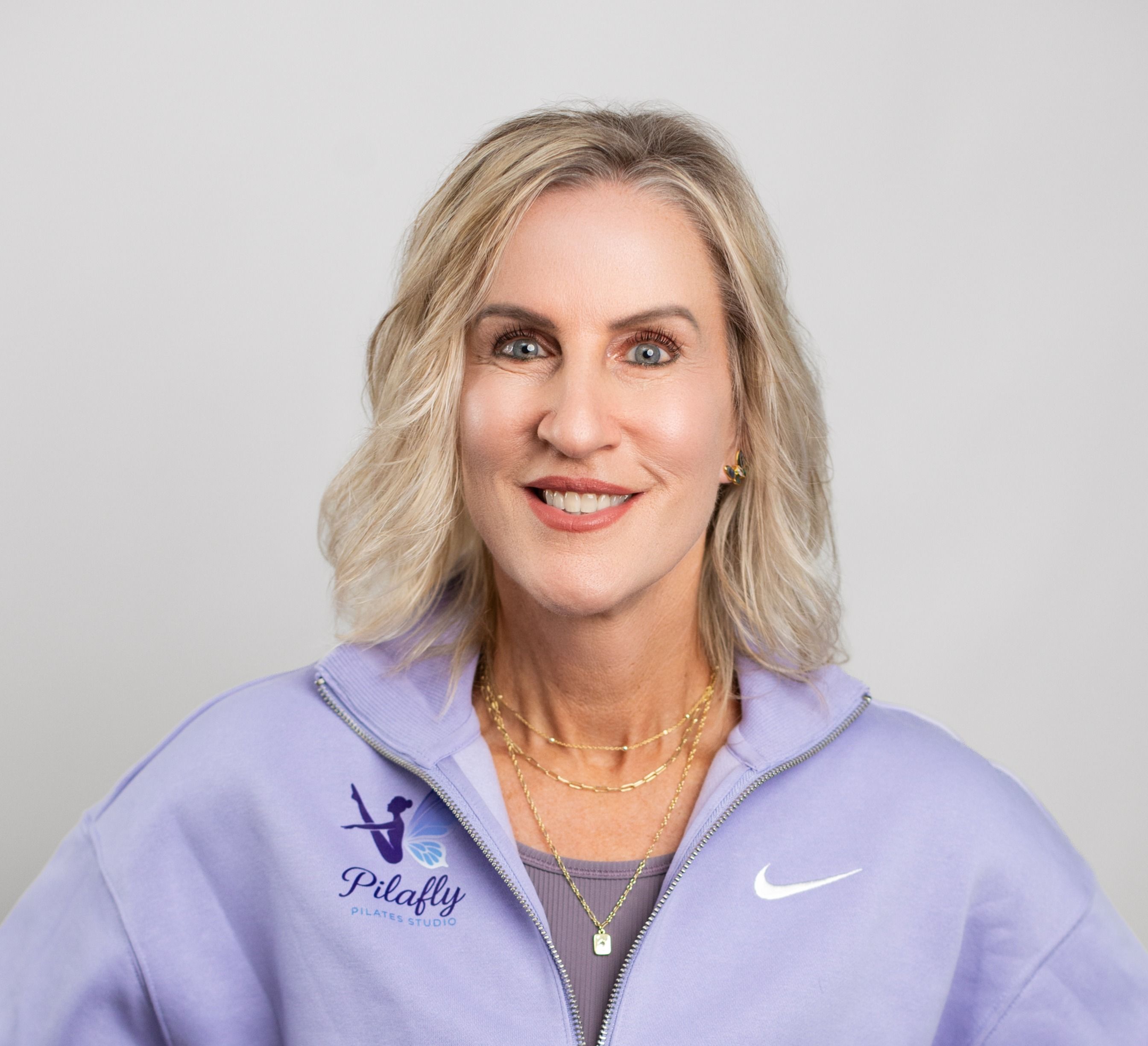 Woman with blonde hair smiling, wearing a purple Nike jacket and gold jewelry, against a plain background.