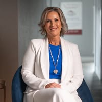 Woman with shoulder-length hair wearing a white blazer and blue top, sitting in an office setting.