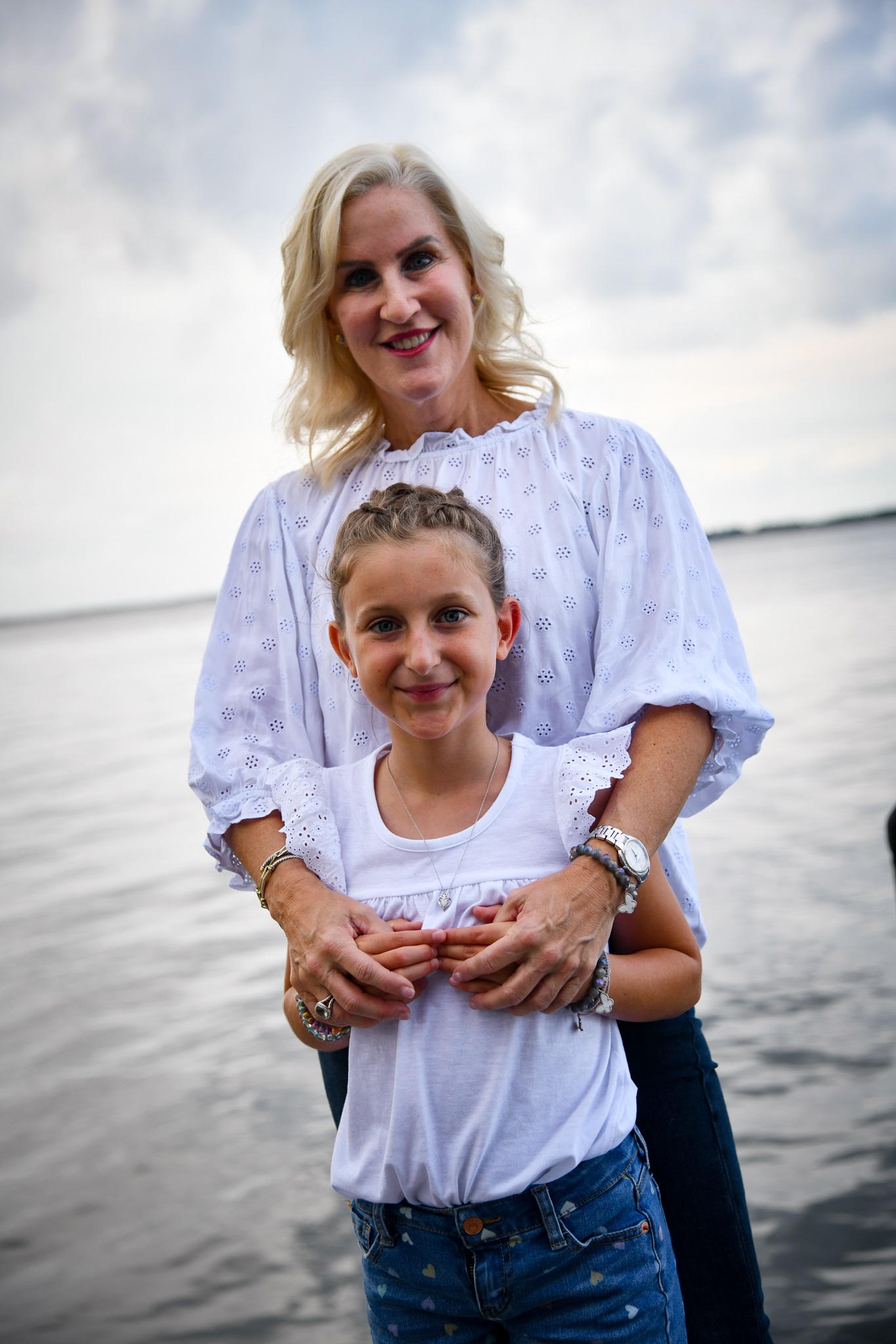 Woman and girl standing outdoors near water, smiling, wearing white tops, cloudy sky background.
