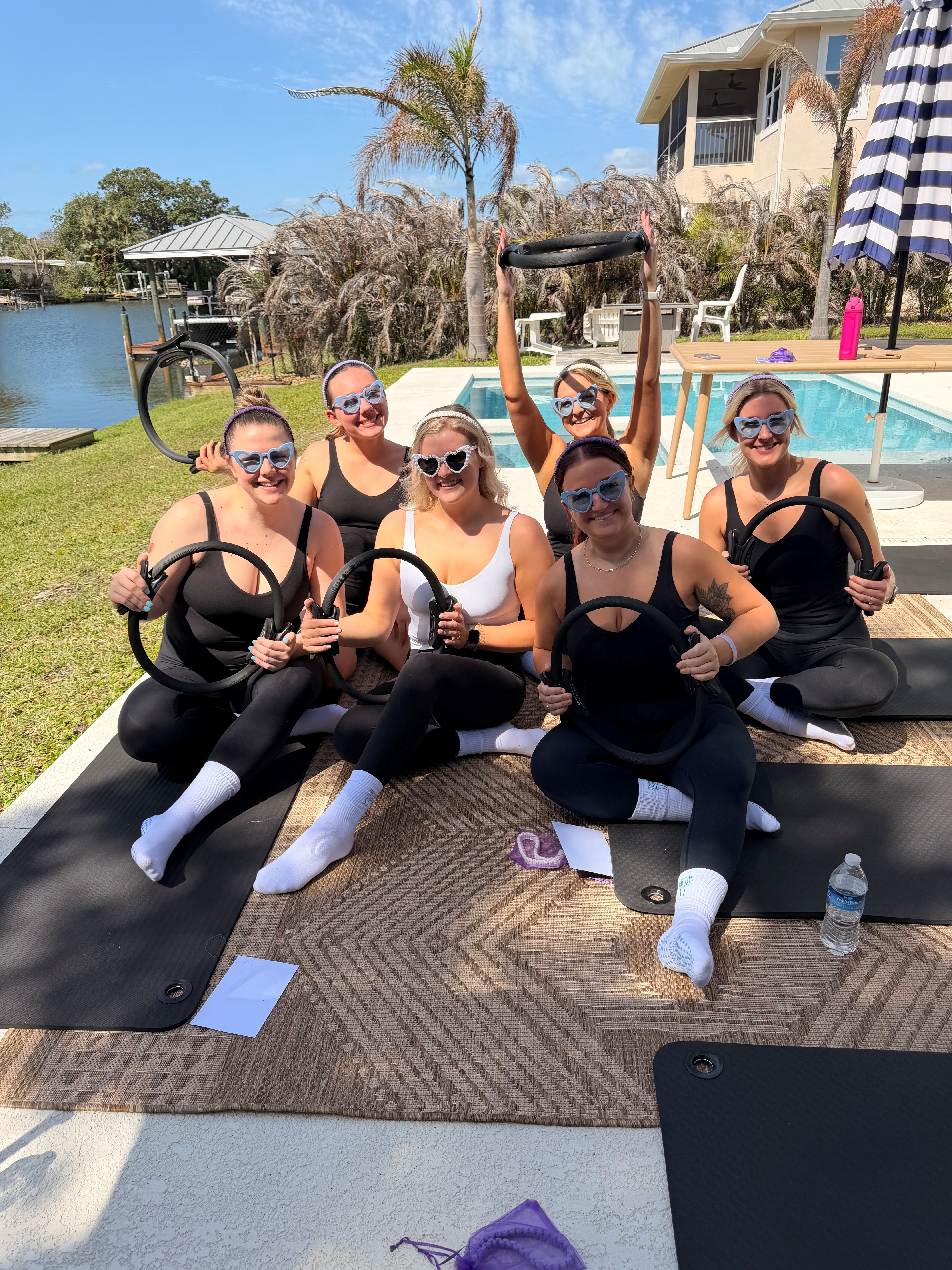 Five women in workout clothes and sunglasses sit on mats outdoors near a pool, holding fitness equipment, smiling.