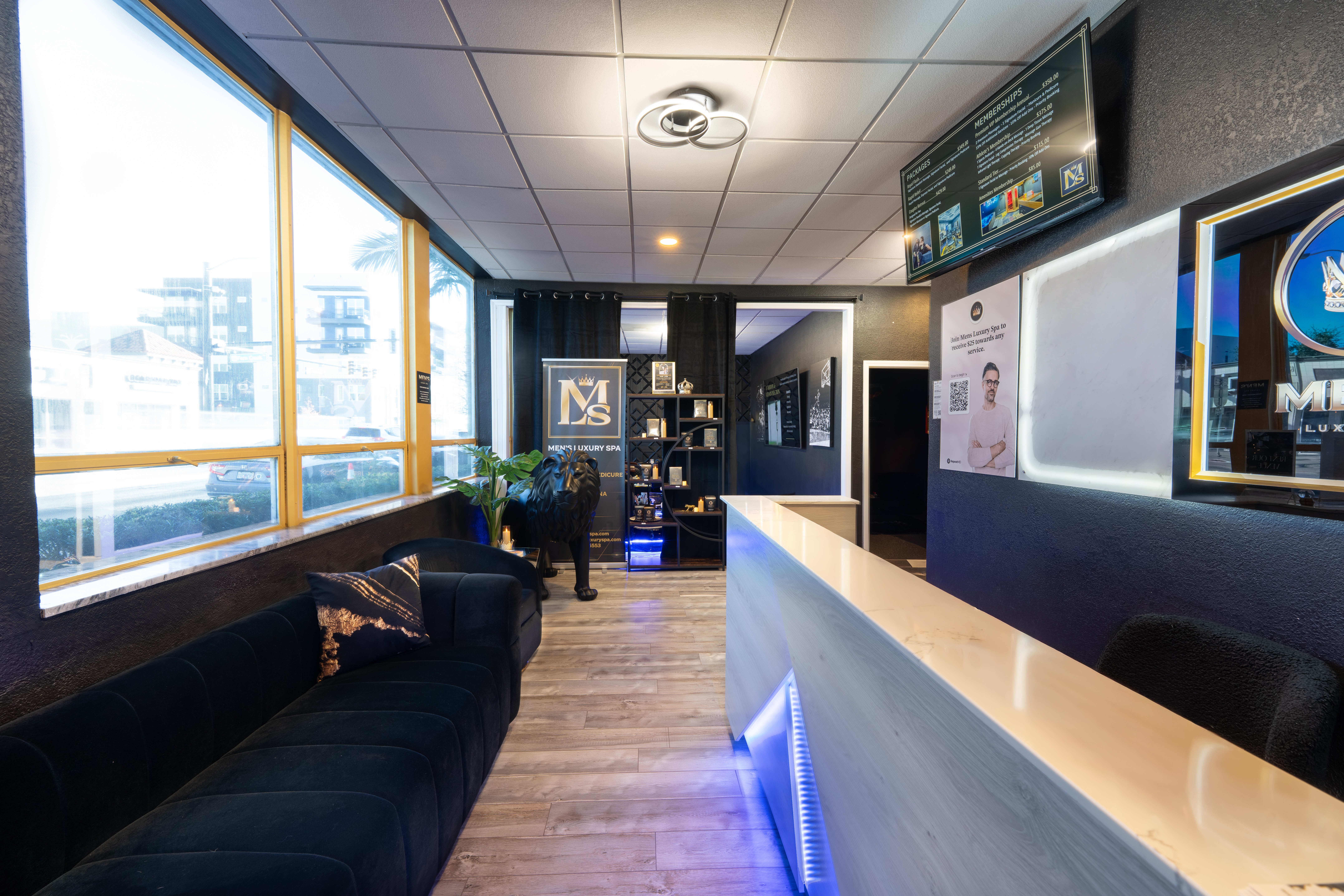 Interior of a modern lounge or reception area with large windows, black seating, and a white counter, illuminated by ceiling lights.
