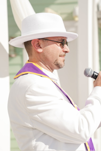 Man in white hat and suit speaking into a microphone outdoors, wearing sunglasses, with a blurred background.