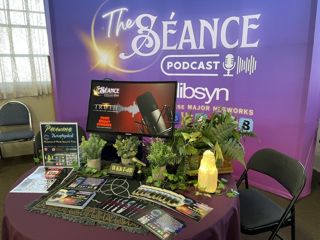 Podcast setup with a table, monitor, microphone, plants, and promotional materials in front of a purple backdrop.