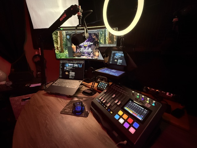 A music or streaming setup with multiple screens, a microphone, a ring light, and a mixing console on a wooden desk.