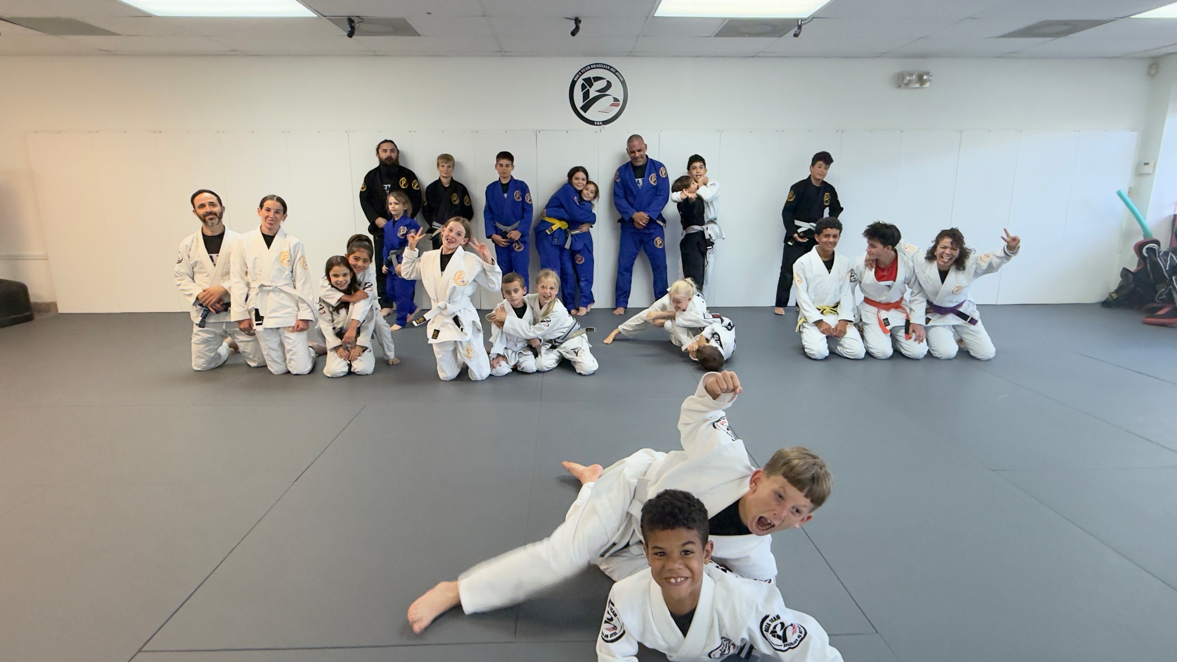 Group of children and adults in martial arts uniforms in a dojo, some sitting and some standing, with a clock on the wall.
