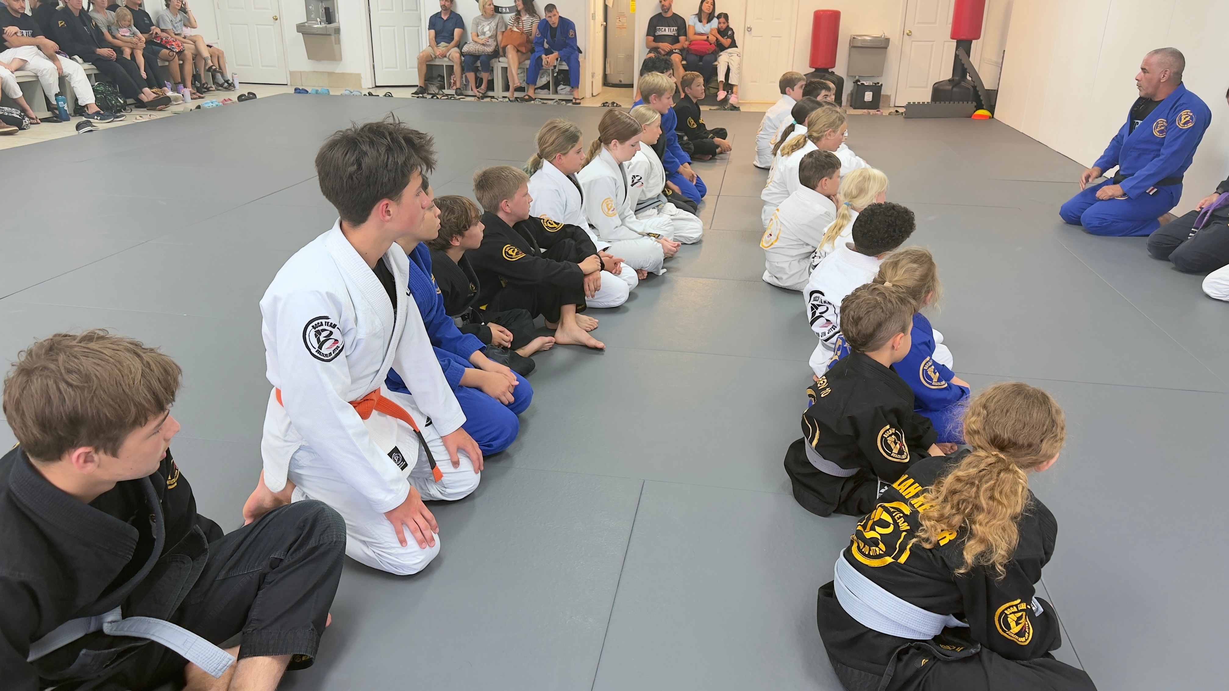 Children sitting in a line on a martial arts mat, listening to instructor in blue uniform, in a training room.